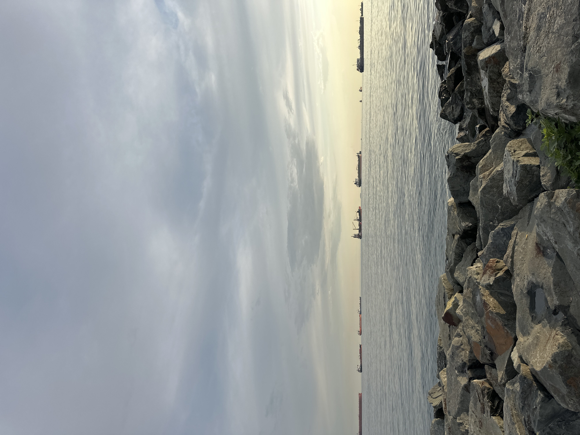 The image depicts a serene coastal scene in Bakırköy, Turkey, overlooking the Marmara Sea. In the foreground, a rocky shoreline composed of large, grey stones leads down to the water's edge. The sea is calm, with gentle ripples reflecting the overcast sky. Several cargo ships are visible in the distance, navigating the expansive waters. The sky is filled with a layer of diffused clouds, with a subtle yellow glow on the horizon suggesting the sun is either rising or setting. There are no people or specific architectural structures immediately apparent in the frame, emphasizing the natural and maritime elements of the landscape.