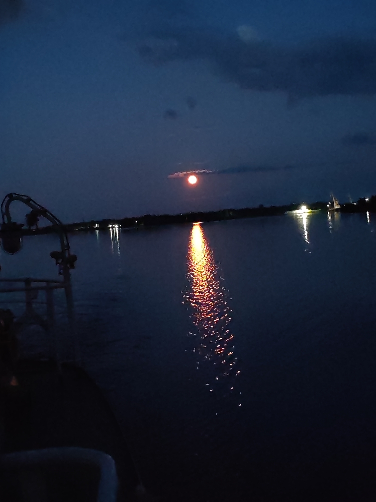 A nighttime view of a body of water, likely a river, with a full moon visible in the dark sky. The moon casts a shimmering reflection onto the water's surface. The foreground shows a portion of a boat or pier with dark railings and supports. In the distance, a dimly lit shoreline is visible with scattered lights, possibly from a settlement. The overall mood is serene and tranquil, characteristic of a calm night. The time of day is clearly night due to the darkness and the presence of the moon. No people are clearly discernible in the image, though their presence is implied by the boat/pier structure. No text is visible.
