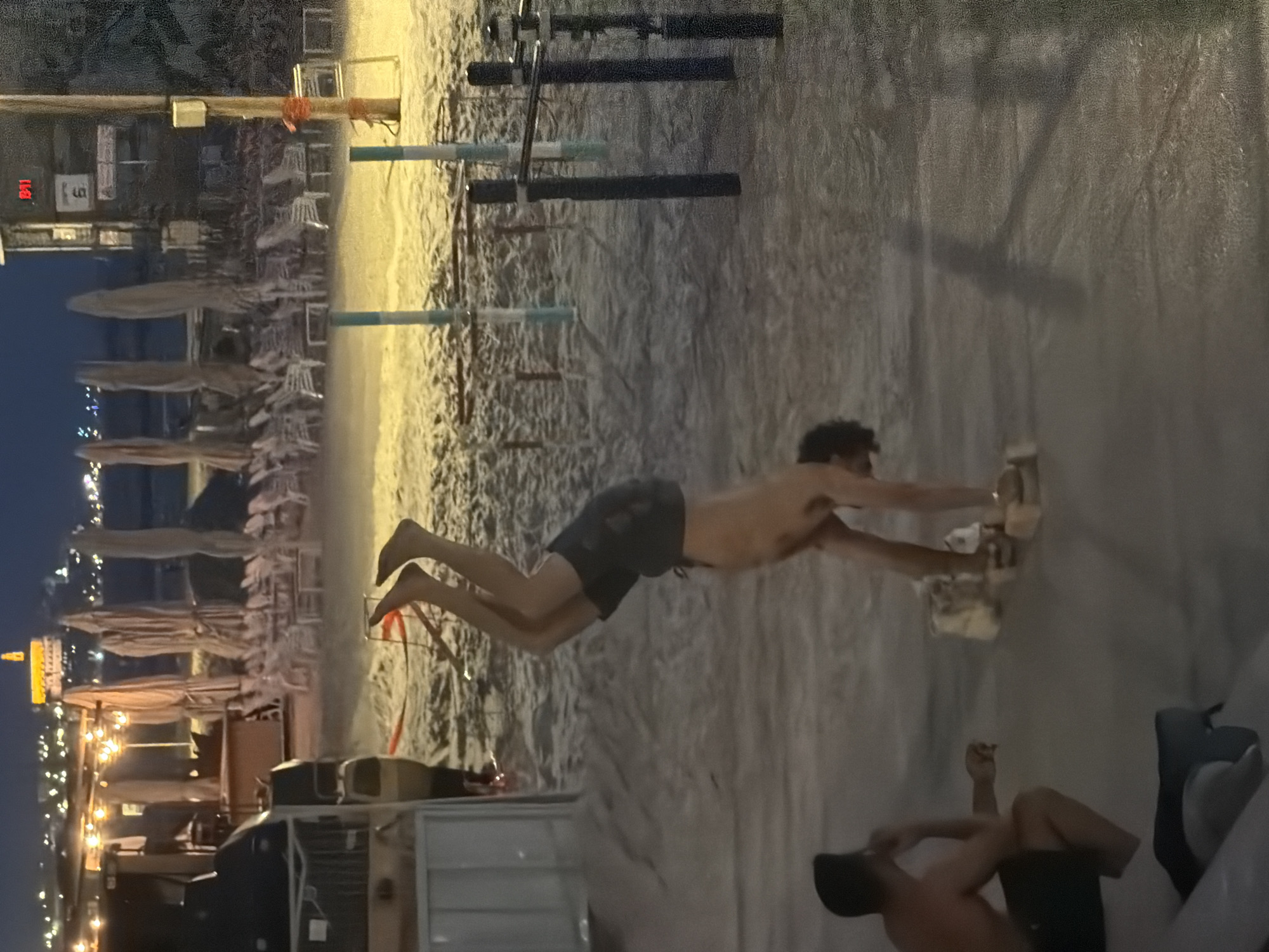 The image depicts a beach scene in Tel-Aviv, Israel, at dusk. In the background, the city skyline is visible with illuminated buildings. Beach umbrellas and chairs are arranged along the sandy beach. In the foreground, two men are present. One man is shirtless and appears to be doing some kind of fitness exercise with weights in his hands while suspended in the air between two supports. Another man is sitting on the ground wearing a hat and shorts. The environment appears to be a combination of sand and paved areas.