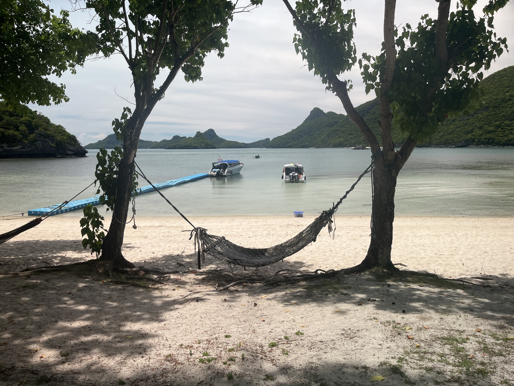 The image shows a tropical beach scene with two trees framing a hammock. The hammock is made of woven rope and is suspended between the trees. The beach is made of white sand, and the water is calm and clear. In the background, there are several islands with lush green vegetation, and two boats are visible in the water. A blue floating dock extends from the shore into the water. The sky is overcast with clouds. The scene appears to be in a serene, natural environment, likely a popular tourist destination in Thailand, such as Ang Thong Marine National Park.