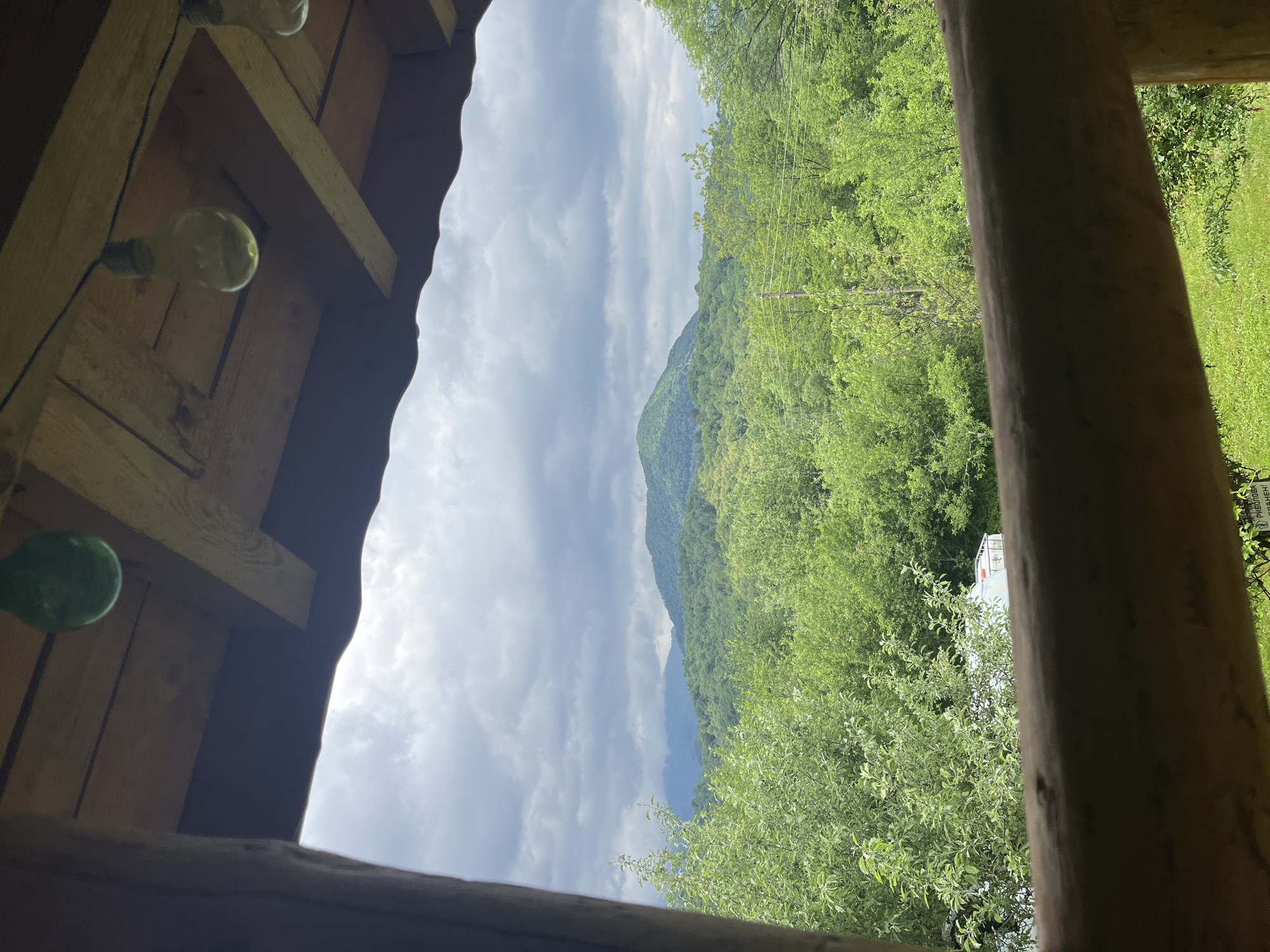 The image shows a view of the sky and surrounding nature, likely taken from inside a wooden structure. In the foreground, sections of a wooden roof and wooden supports are visible, with light bulbs hanging from the roof's interior. Through the opening in the structure, a landscape unfolds, featuring a cloudy sky and a mountain range covered in dense green foliage. The scene could be located in a natural setting near Shopite, Bulgaria, as indicated by the context. The presence of a house among the trees suggests a semi-rural or suburban environment. No people or specific actions are discernible. The scene is predominantly natural, with the human-made structure providing a frame for the landscape view.