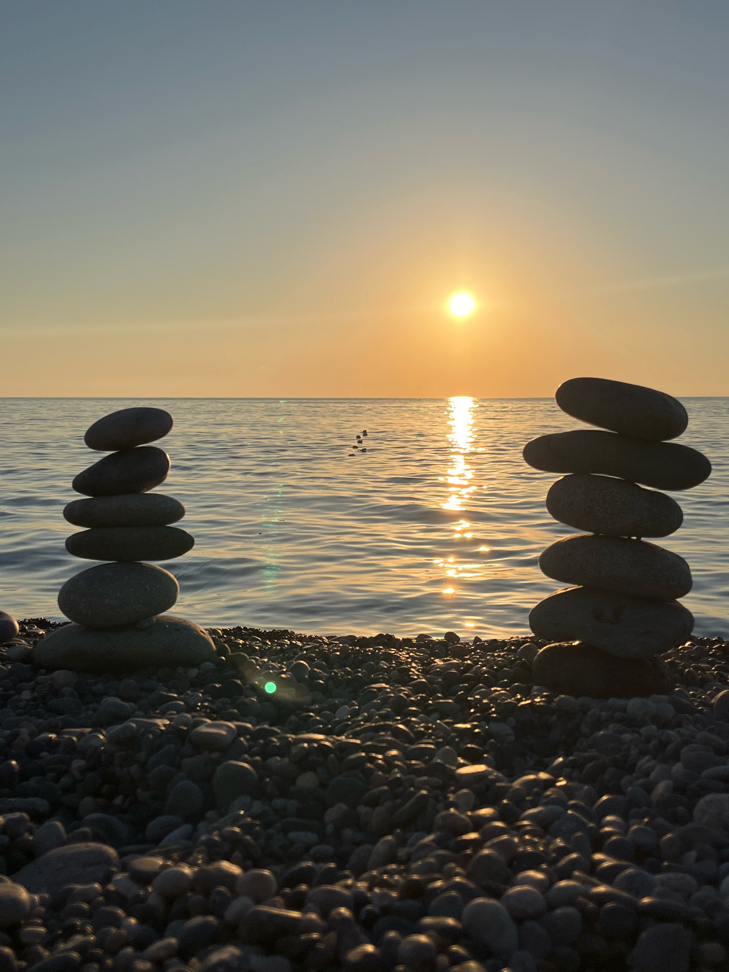 The image shows a tranquil scene on a pebble beach at sunset in Kobuleti, Georgia. Two stacks of flat, smooth stones are balanced on top of each other, with the one on the left having five stones and the one on the right having six. The sun is low on the horizon, casting a warm, golden glow across the calm sea, which reflects the light in a shimmering path. The sky is a gradient of soft blue and orange. The foreground is filled with an abundance of dark gray and black pebbles, some catching the sunlight. There are no people visible in the scene, and the overall impression is one of peaceful solitude and natural beauty.