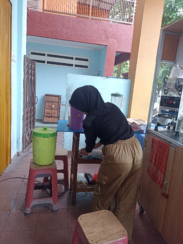 This medium shot captures a person, viewed from the back, actively engaged in what appears to be food or drink preparation at an outdoor or semi-outdoor stall in Tulungagung, Indonesia. The individual wears a black hijab, a black long-sleeved top, and loose-fitting khaki cargo pants, bending over a simple wooden table.
On the table, a purple jug is visible, and behind it, a blender suggests the preparation of beverages. Below the table, various items like a black remote and a red object are arranged on a lower shelf. To the far left, a bright green water dispenser with a lid rests on a red plastic stool, further indicating a drink preparation or serving station. Adjacent to the person on the right is a mobile stall equipped with what appears to be a cup sealer or similar beverage preparation machinery, with a red checkered towel hanging on its side.
The setting features a tiled floor and light blue walls in the background. An upper level of a building with a reddish-brown facade and a white railing is also visible. Natural, bright daylight illuminates the scene, suggesting it is daytime. On a yellow vertical surface to the left, a small, white sign with indistinct text is present. The overall environment depicts a typical small-scale vendor setup in Southeast Asia.