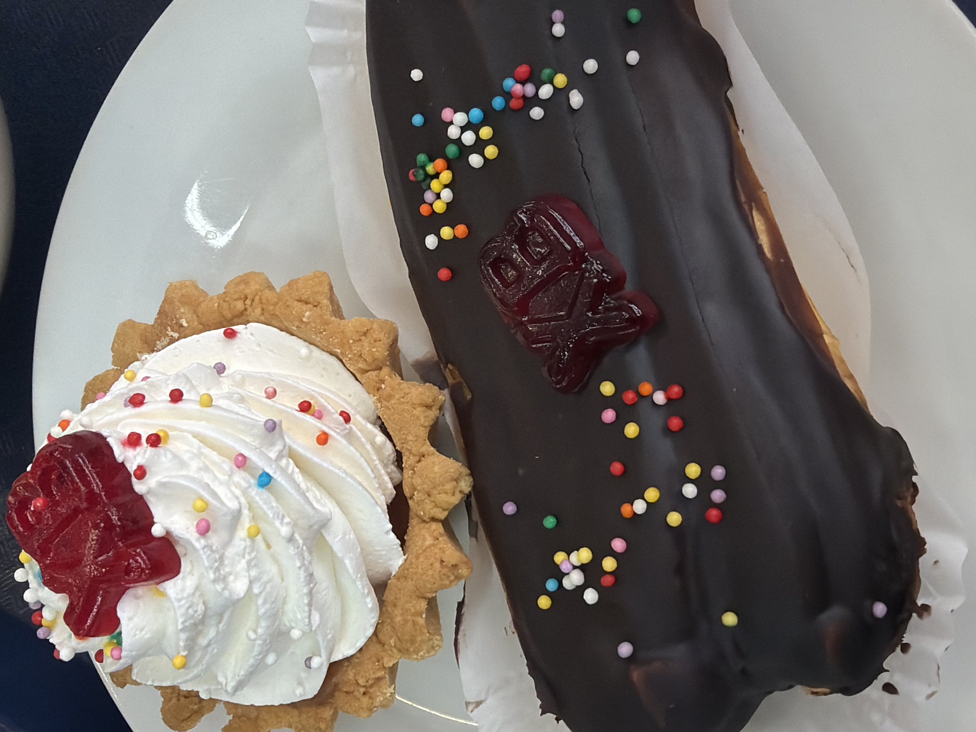 A white plate contains two pastries: a tartlet filled with white whipped cream and topped with a red jelly candy, and a chocolate-covered eclair also topped with a red jelly candy. Both pastries are decorated with small colored sprinkles. The pastries appear to be photographed in Perm, Russia.