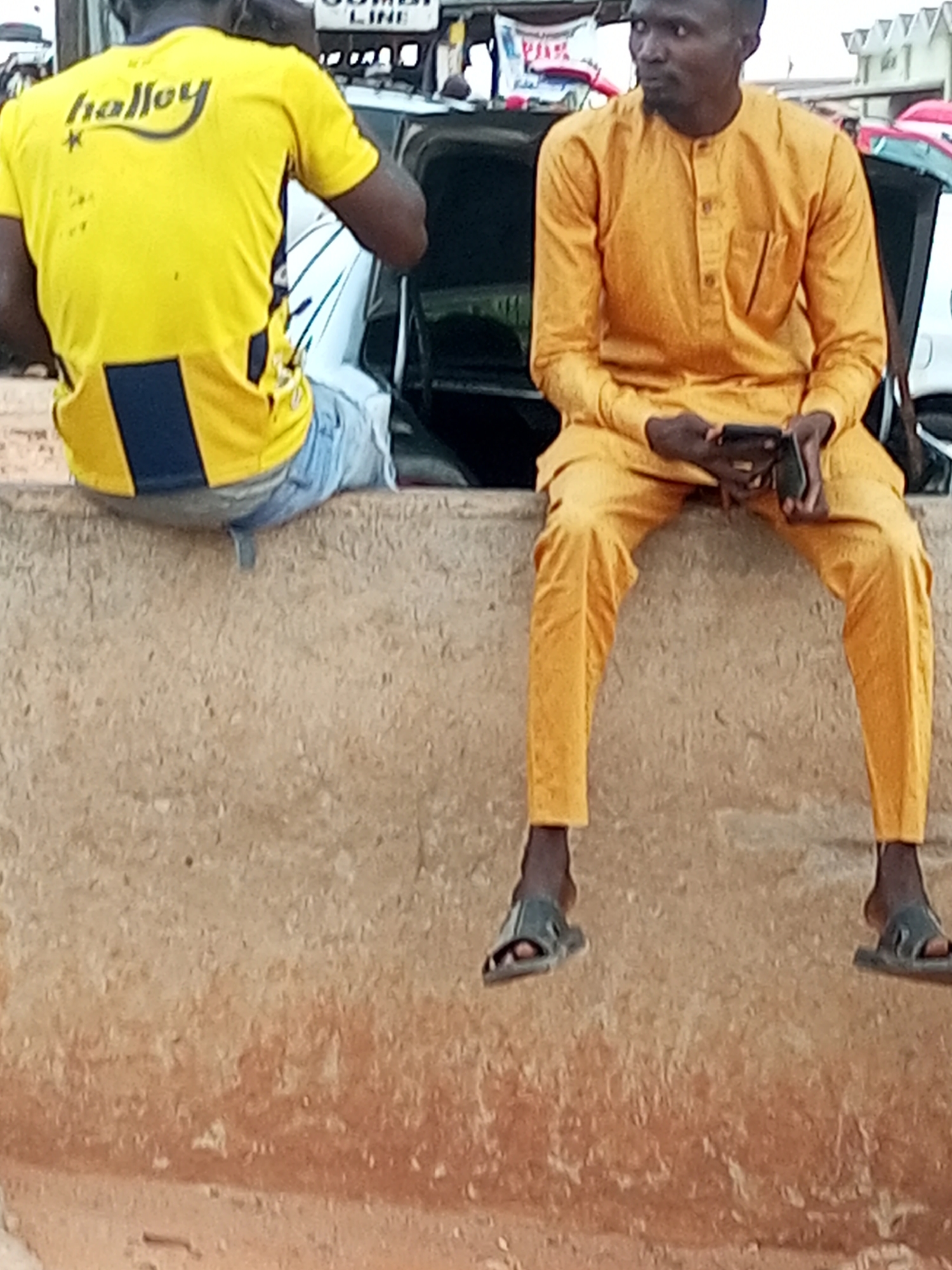 Two individuals are seated on a concrete ledge. The individual on the left is wearing a bright yellow t-shirt with "halloy" written in black script on the back and a dark blue rectangular panel on the lower back. They are also wearing light blue jeans. The individual on the right is wearing a mustard-yellow, long-sleeved tunic with buttons down the front, and matching yellow trousers. They are holding a dark mobile phone in their right hand and have a black bag slung over their left shoulder. Both individuals are wearing dark-colored sandals. The background suggests an urban setting in Gombe, Nigeria, with parts of vehicles and some signage visible, indicating a possible transportation hub or marketplace. The overall environment appears to be outdoors, with a textured concrete wall in the foreground.