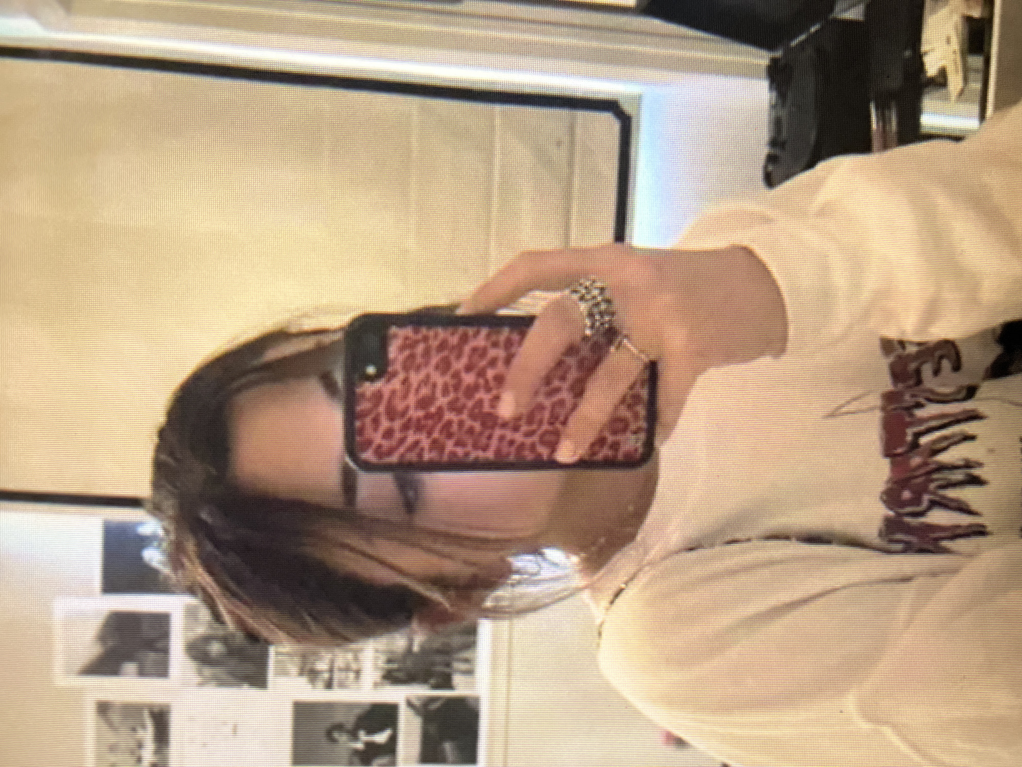 A young woman is captured in a slightly blurred, low-resolution selfie taken in a room. She is holding up a smartphone with a red leopard-print case, angled to show her reflection in a mirror that is not fully visible. Her hair is pulled back, and she is wearing a light-colored long-sleeved shirt with a graphic print on the front. She has multiple rings on her fingers. Behind her, the wall is decorated with several small black and white photographs. The lighting in the room appears to be natural, casting a warm glow. The environment suggests a personal space, possibly a bedroom or dressing area. There is no indication of the specific location in the city of Qarağandı city administration, Kazakhstan.