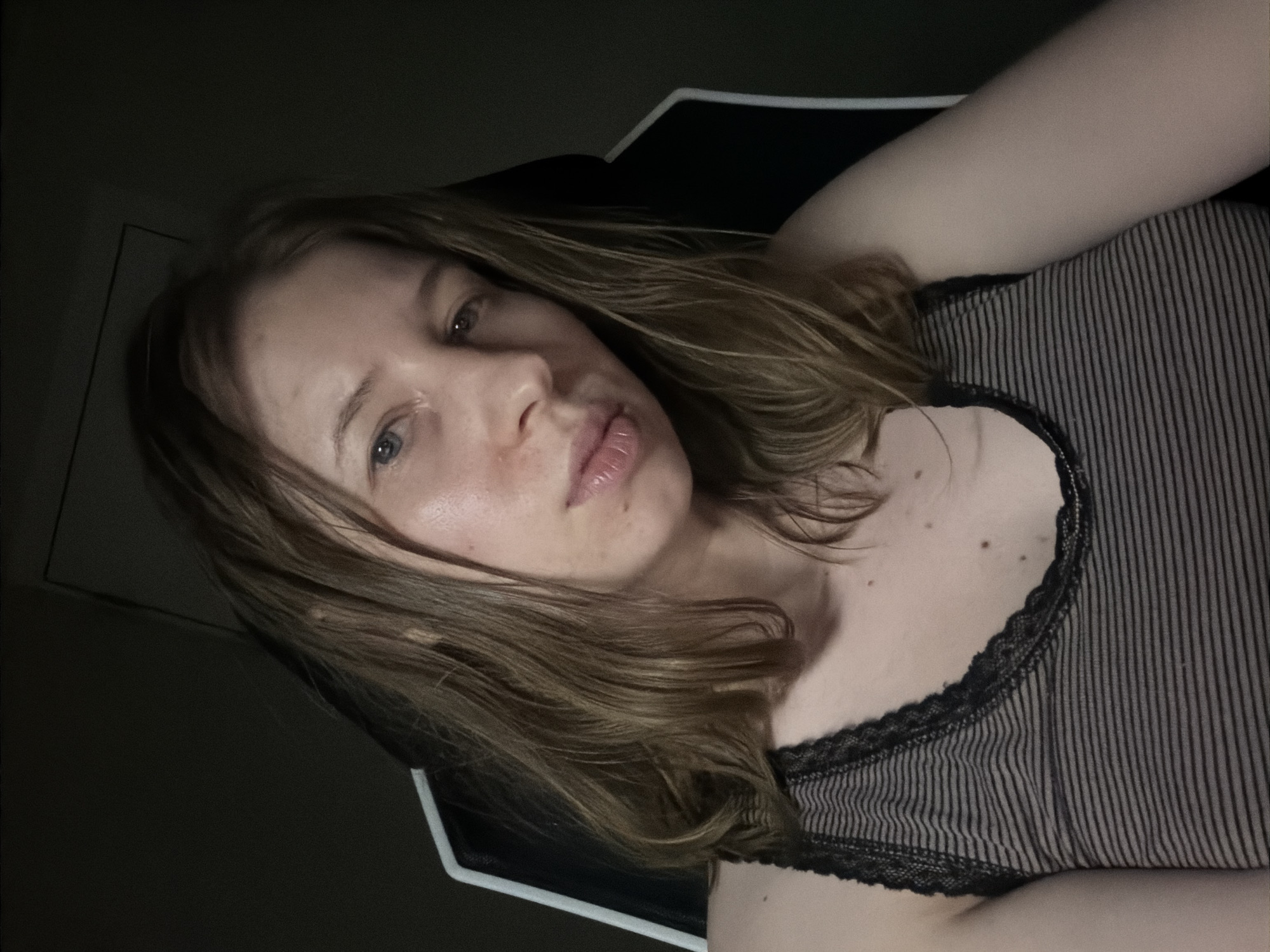 A young woman with shoulder-length, wavy blonde hair is pictured in a dimly lit room, seated in what appears to be a gaming chair. She is looking directly at the camera. She is wearing a dark-striped tank top with black lace trim. The background is dark and indistinct, offering no specific environmental clues or details about the location in Dongducheon-si, South Korea.