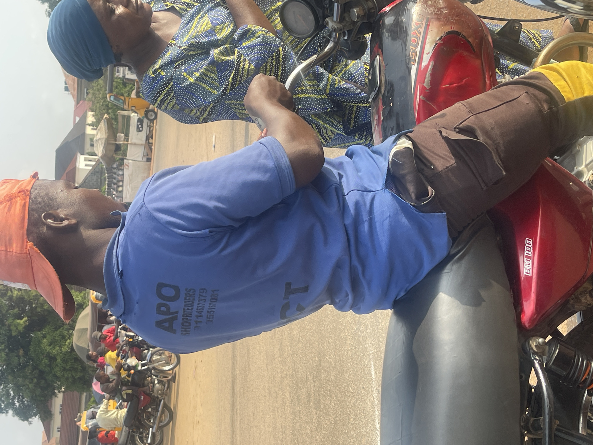 This image captures a scene in Galadimawa Village, Nigeria. Two individuals are prominently featured, seated on a motorcycle. The person in the foreground, wearing an orange cap and a blue shirt, is looking to the side. Their shirt displays text that reads "APO SHOPPRENDERS" followed by a series of numbers: "01146379", "355167031". The second person, seated behind, is wearing a blue headscarf and a blue and yellow patterned traditional garment. The motorcycle is red and black.
In the background, a row of other motorcycles is visible, suggesting a gathering or transport hub. A yellow kiosk-like structure and some umbrellas are also present, indicating a market or roadside vendor area. The overall impression is that of a typical day in a Nigerian village, likely during daylight hours, with a warm, possibly sunny, atmosphere. The image appears to be taken from a slightly elevated angle, looking down at the subjects and the surrounding scene. The focus is primarily on the two individuals and the motorcycle they are on, with the background providing context to their location and the potential activities occurring around them.