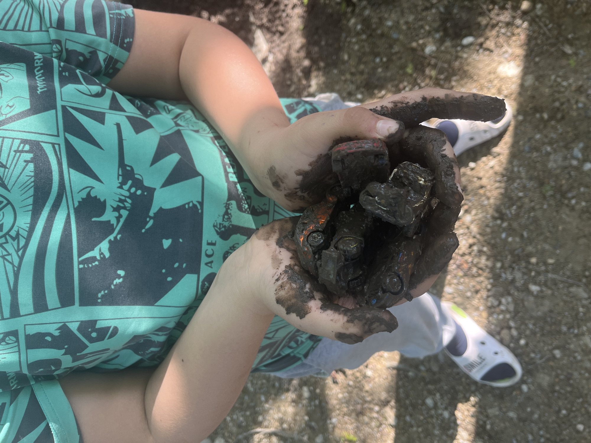 A child is holding a cluster of toy cars covered in mud in their hands. The child is wearing a teal t-shirt with a comic-book style pattern and grey sweatpants. The child is also wearing white Crocs with blue accents. The background is out of focus and shows what appears to be a gravel surface. The city location is unknown.