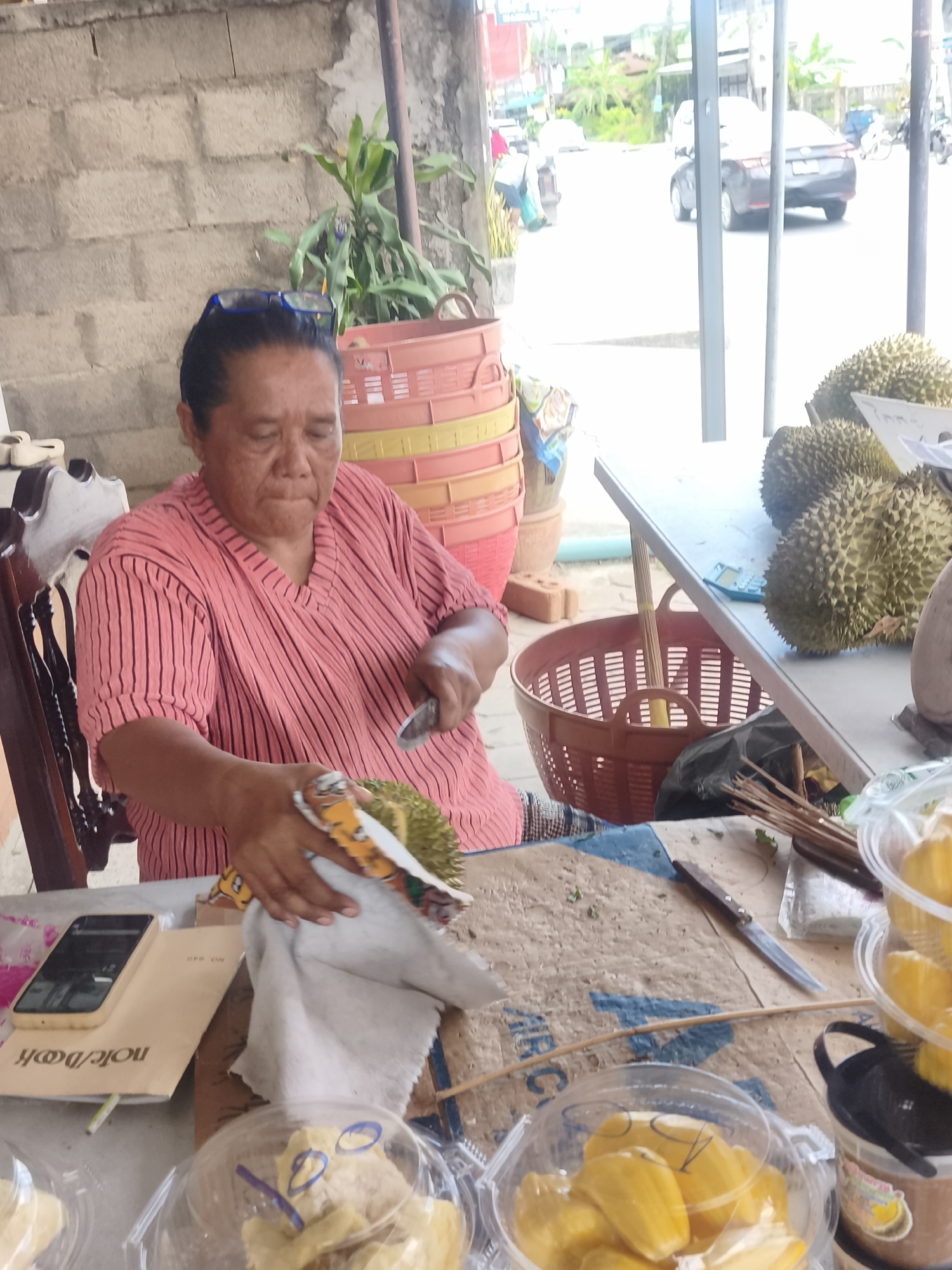 In an outdoor market setting, a woman with dark hair and glasses, wearing a pink striped shirt, is seated and preparing a durian fruit. She is holding a knife in her right hand and a piece of cloth in her left, with the durian partially peeled. Several other durian fruits are visible on a table to her right, along with plastic containers filled with yellow fruit pieces, possibly jackfruit. In the background, stacked baskets are visible, and beyond them, a street scene with vehicles and buildings can be seen. The ground is covered with a rough material, and a mobile phone and notebook are on the table in front of the woman.