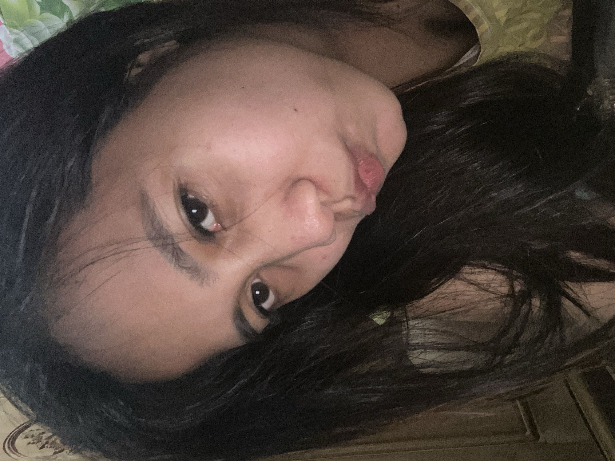 A close-up, head and shoulders shot shows a young woman with long, dark hair, slightly angled towards the right of the frame. Her eyes are dark and looking towards the left, with a slight pout to her lips. The lighting appears to be indoor, with a warm tone, suggesting it was taken in a residential setting. A portion of a patterned wall or fabric is visible in the background. The image does not provide enough detail to definitively place it within the city of Depok, Indonesia.