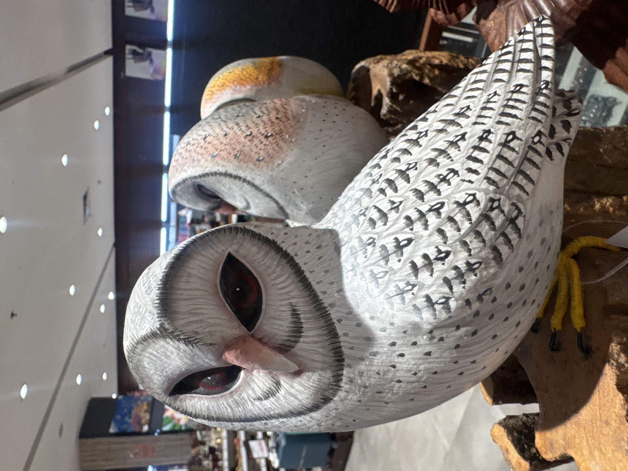 The image displays a close-up of a white owl figurine with striking red eyes and a pink beak, positioned on a wooden base. The owl's feathers are intricately detailed with gray markings. To the right of the main owl, two other owl figurines are partially visible. The one immediately behind has a white body with subtle orange and brown speckles on its head, while the furthest one is mostly out of focus with a yellow and orange head. The background suggests an indoor setting, possibly a shop or market, with displays and shelving visible. The lighting appears to be from overhead fluorescent lights. The overall scene depicts decorative owl figures arranged for display.