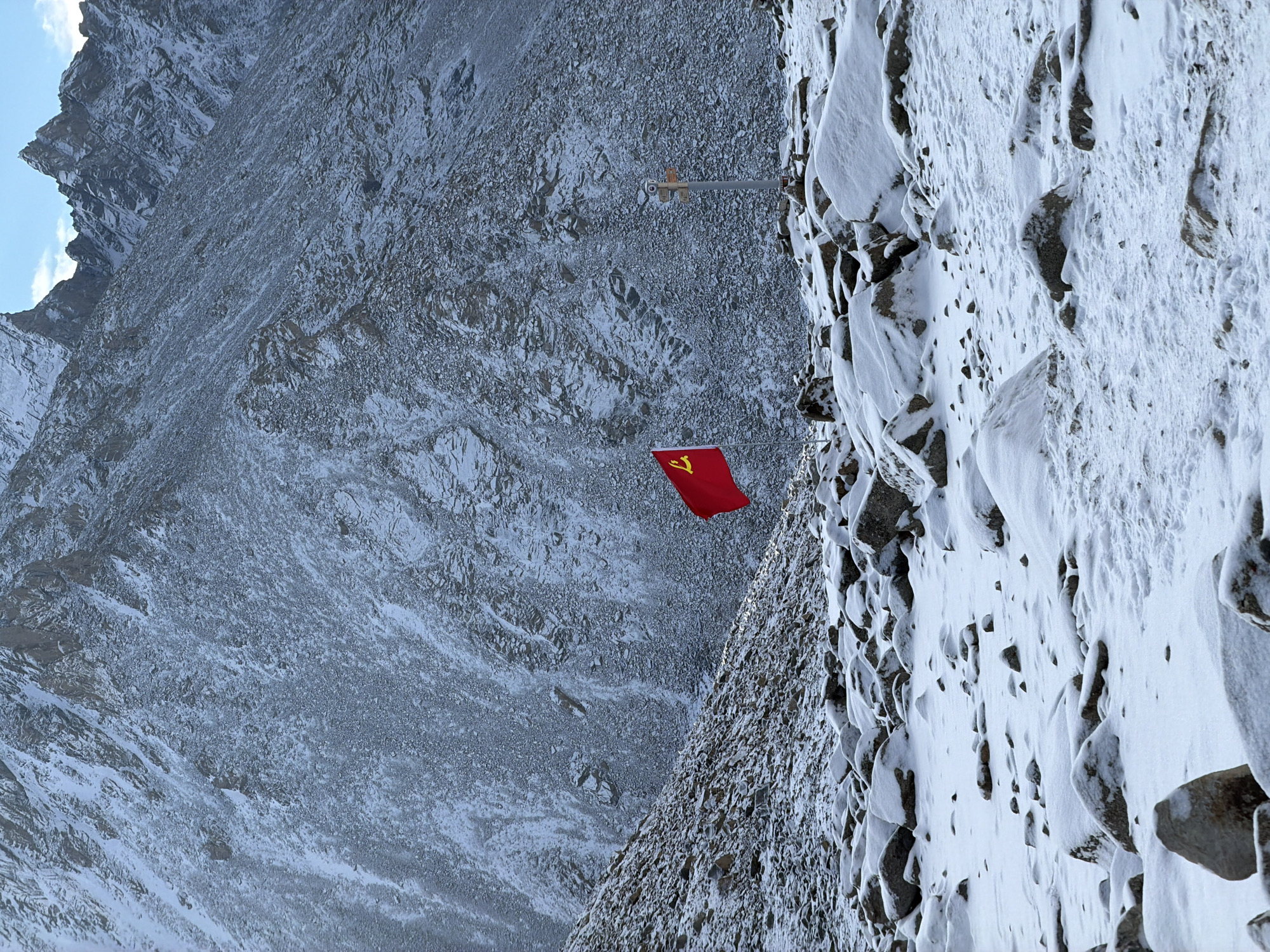 The image displays a red flag adorned with a yellow hammer and sickle symbol, firmly planted on a steep, snow-covered mountain slope. The flag is positioned on what appears to be a metal flagpole secured to the rocky terrain. The surrounding landscape is rugged, characterized by jagged peaks and extensive rock formations, all dusted with a layer of snow.
The scene depicts a harsh, high-altitude environment, likely a mountain range, under a clear daytime sky with sparse clouds visible in the upper left background. The ground is a mix of exposed dark rock and white snow, suggesting cold, wintry conditions. There is no human activity explicitly visible, but the presence of the flag indicates human placement. The flag itself is the most prominent man-made object, flying in the wind against the backdrop of the desolate mountains. The hammer and sickle emblem is a notable detail, historically associated with communist parties, and in this context, strongly suggests the flag of the Communist Party of China given the provided location context. The overall impression is one of remoteness and resilience, with the flag serving as a prominent marker in a challenging natural setting. No other visible text is present.