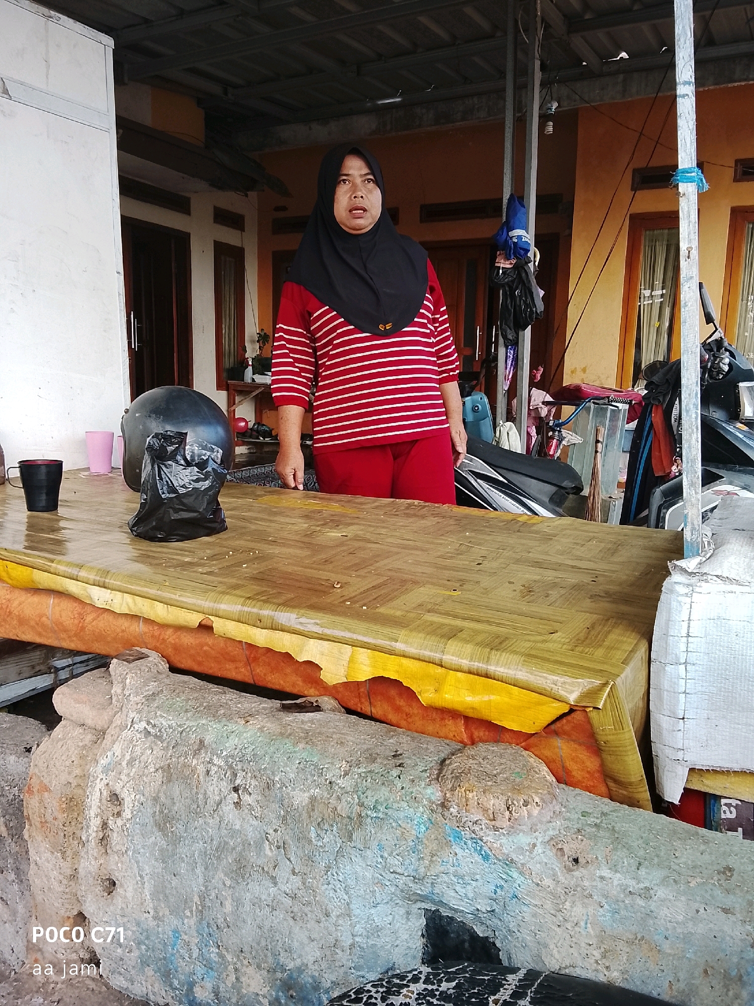 A woman wearing a red and white striped long-sleeved shirt and red pants, covered by a black hijab, stands behind a table covered with a yellow plastic sheet. Her facial expression appears surprised or concerned. On the table, there is a black helmet with a black plastic bag on top of it, a black mug, and two pink cups. The table is supported by a structure that appears to be made of concrete or stone, with visible rough textures and colors. In the background, there is a house with orange walls and wooden doors and windows. There are also several motorcycles and other items that suggest a marketplace or a roadside stall. The scene is outdoors, under a metal roof supported by posts. The lighting suggests it is daytime. The bottom left corner of the image displays "POCO C71" and "aa jami" which are likely camera model and watermark information.