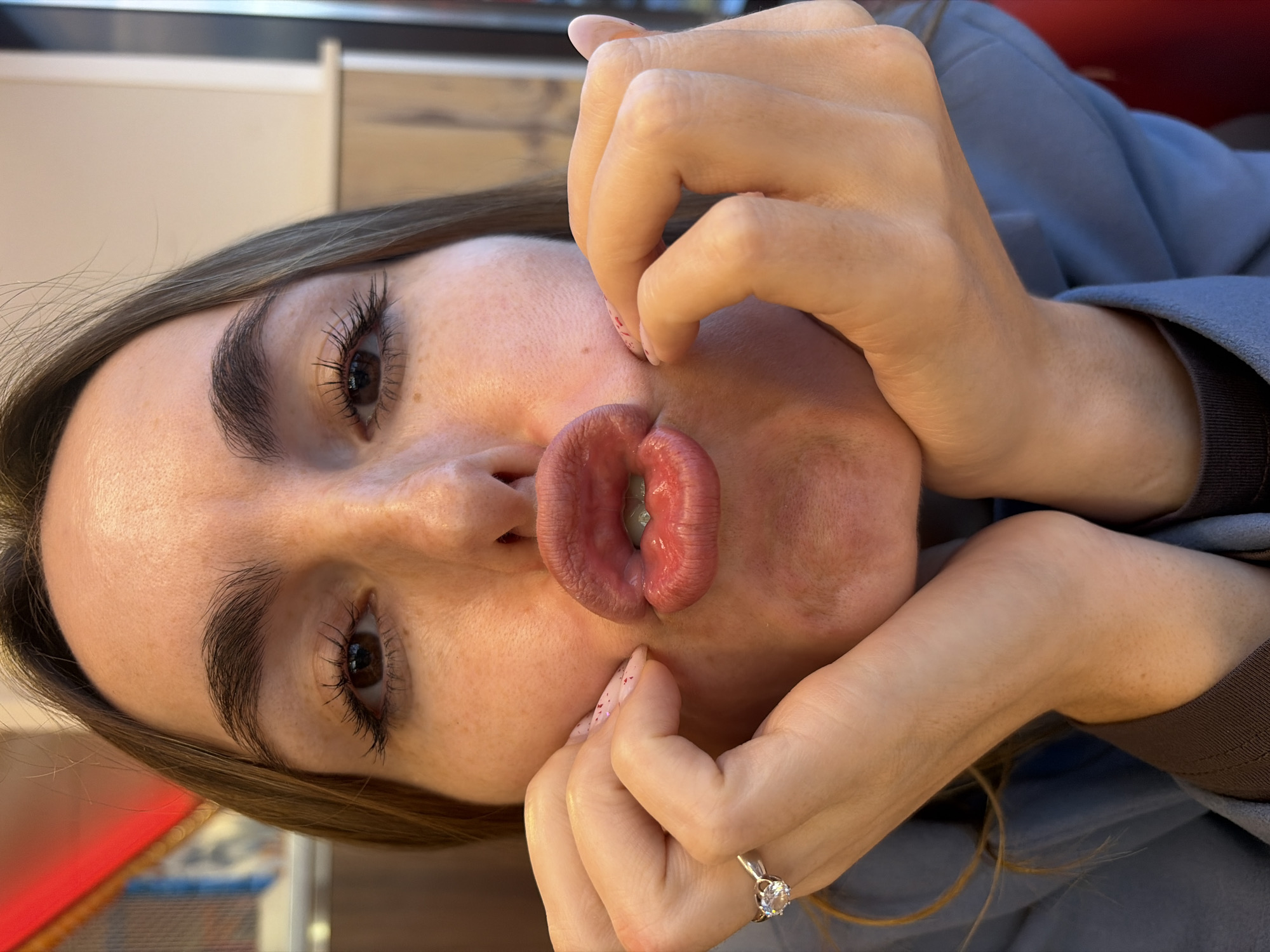 A female individual is depicted in a close-up, head-and-shoulders portrait shot, taken indoors in Izhevsk, Russia. She faces forward, making a pronounced "pouting" or "fish face" expression with her lips tightly pursed and pushed forward. Both of her hands are raised, with fingers pressing into her cheeks, further accentuating the facial distortion.
The individual has medium brown hair, brown eyes, and dark, well-defined eyebrows. Her eyelashes are prominent, suggesting mascara application. Freckles are visible across her nose and upper cheeks. She is wearing a light blue outer garment over a dark brown long-sleeved top. A silver-toned ring with a clear, faceted stone is present on the ring finger of her left hand. Her fingernails are coated with a light pink polish speckled with small, reddish glitter particles.
The background is out of focus, indicating an indoor commercial environment, possibly a cafe or restaurant. Visible elements include an indistinct bright red object on the left, and lighter-toned walls with vertical light wooden trim or paneling on the right. The scene is evenly lit by artificial light.