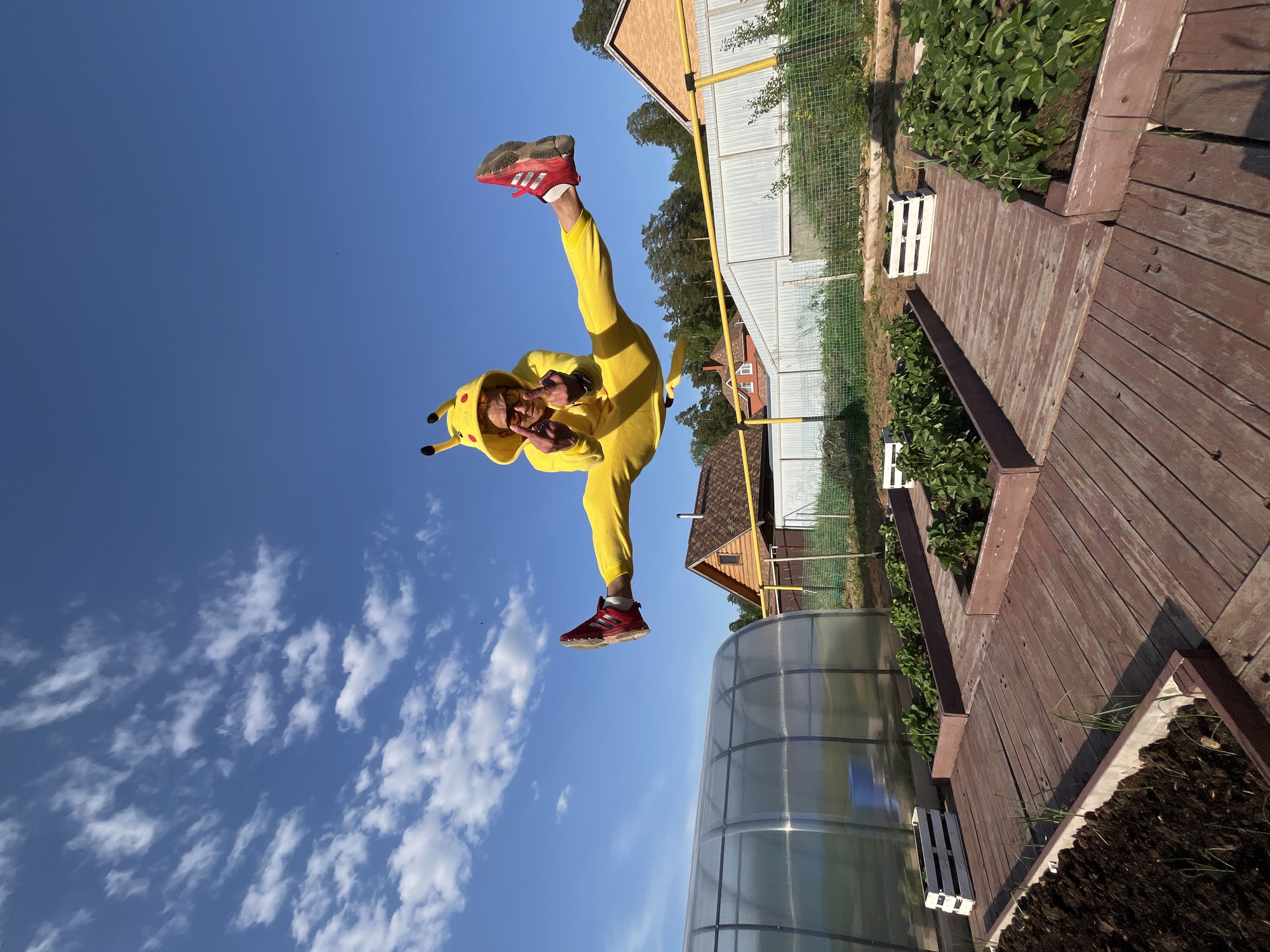 A person in a Pikachu costume is captured mid-air against a backdrop of a clear blue sky with scattered clouds. The individual is wearing red sneakers and is oriented upside down. The location appears to be a backyard in Chaikovsky, Russia, featuring a garden, a greenhouse with a translucent roof, a wooden walkway, and residential buildings in the background. A yellow support structure is visible along with wire fencing.
