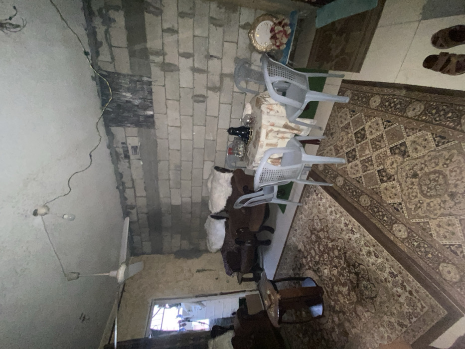 The room has brick walls, and a rug covers the floor. A table with a patterned cloth and decorative items is surrounded by plastic chairs. A brown couch with white covers is adjacent to a small wooden table. A ceiling fan hangs from the ceiling. A window provides natural light into the room.