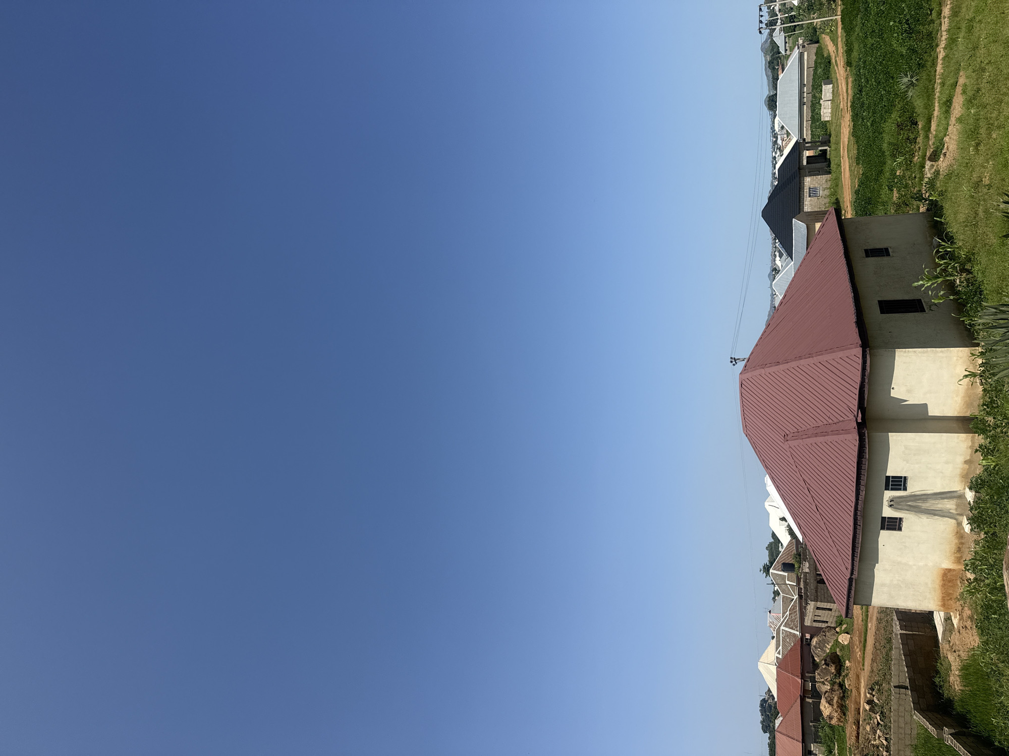 This is a photograph of a residential area under a clear blue sky in an unspecified location. In the foreground, a light-colored, single-story house with a dark red, gabled roof dominates the scene. Two rectangular windows are visible on its facade, and a net is draped over the exterior wall below one of them. Green vegetation, including tall grass and some crops, surrounds the house.
Further in the background, a cluster of other houses with various roof styles, including gabled, flat, and conical, are visible. Some of these homes appear to be under construction, with exposed brickwork and building materials. The landscape is somewhat uneven, with mounds of earth and large rocks scattered in some areas. Power lines and utility poles are also present, indicating the presence of infrastructure. The overall impression is of a developing suburban or rural community.