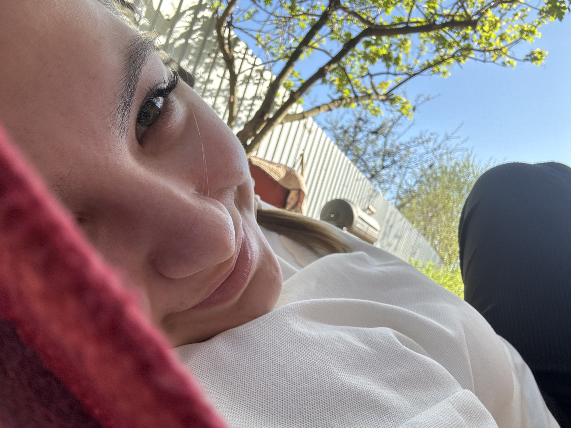 A close-up, low-angle shot captures a person lying down, seemingly relaxed. The subject is fair-skinned with brown eyes and long eyelashes. Their face occupies the left side of the frame, with a strand of hair visible near their nose. They are resting on a white, textured garment. To the right of the subject, a portion of a black garment is visible.
Behind the subject and to the upper right, a glimpse of outdoor scenery is present. A light blue sky is partially obscured by tree branches with light green leaves. The background includes a white, corrugated metal fence and what appears to be a piece of outdoor equipment.
Given the request for a location in Prikamskii, Russia, without additional context within the image, it cannot be verified this image was taken there. This analysis assumes it's a generic outdoor scene possibly associated with the location. There are no identifiable landmarks, signs, or specific architectural features to directly confirm the city of Prikamskii in Russia.