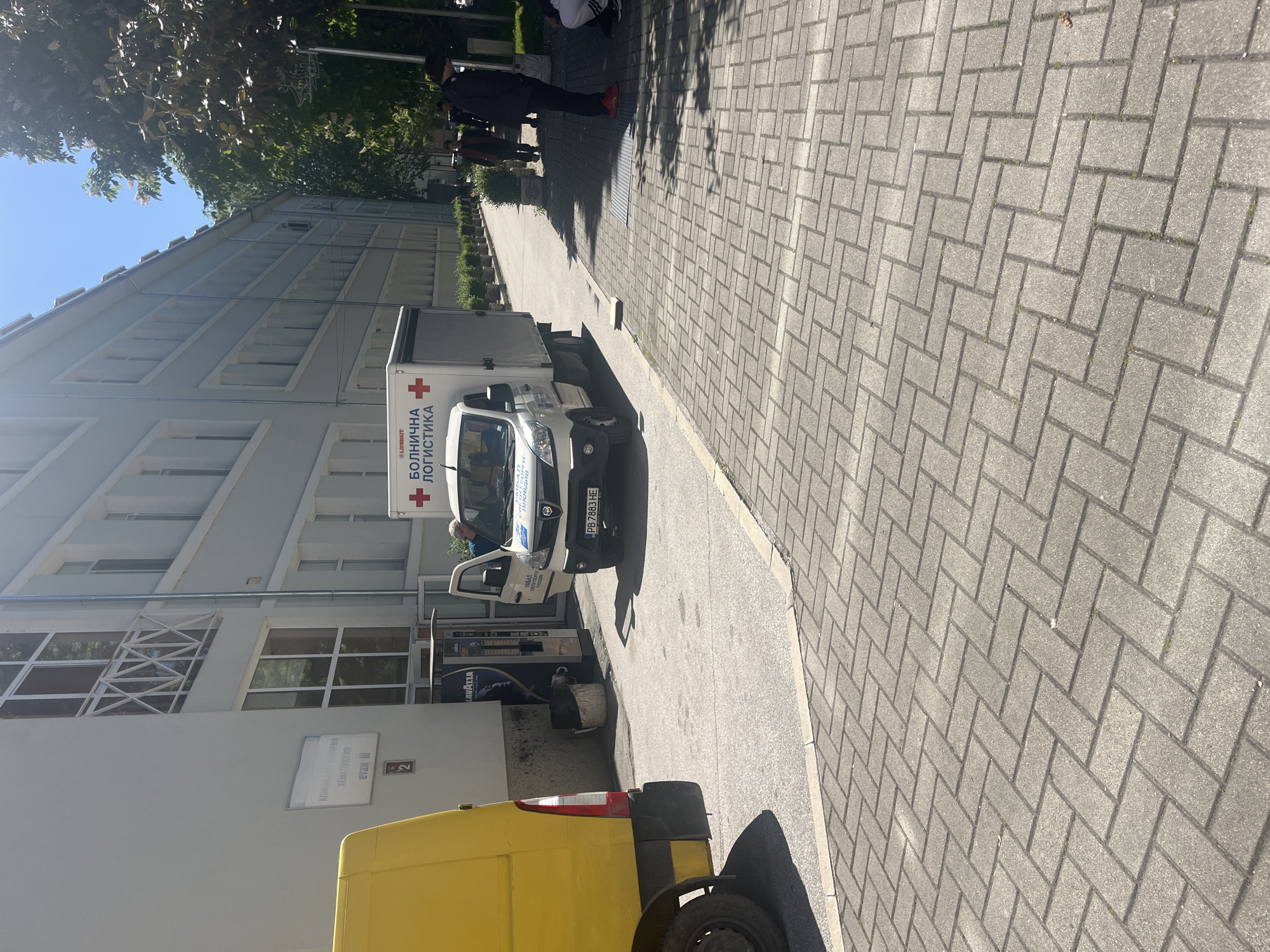 Here is a description of the image:
The image shows a daytime scene outside a building in Plovdiv, Bulgaria. A white van with the words "БОЛНИЧНА ЛОГИСТИКА" (Hospital Logistics) and a red cross is parked near the entrance. A sign on the building reads "КЛИНИКА ПО ВЪТРЕШНА ХЕМАТОЛОГИЯ, етаж 2" (Clinic of Internal Hematology, floor 2). A yellow van is partially visible in the foreground. There are people walking on a paved sidewalk. The license plate of the white van is "PB 7883 HE".