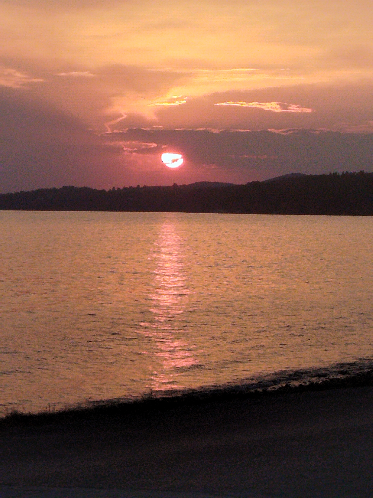 The image shows a sunset over a body of water, possibly a lake or the sea, with a silhouetted treeline and hills in the background. The sky is filled with hues of orange, pink, and purple, with scattered clouds. The sun, partially obscured by clouds, casts a bright reflection on the water's surface. A dark, sloped foreground suggests a shoreline or embankment. The scene appears to be a natural landscape, likely in a rural or coastal setting. While the image is of Greece, the exact location within Sithonia Municipal Unit cannot be determined without further information. There are no people or specific man-made objects visible in the frame, emphasizing a serene and natural environment during twilight.