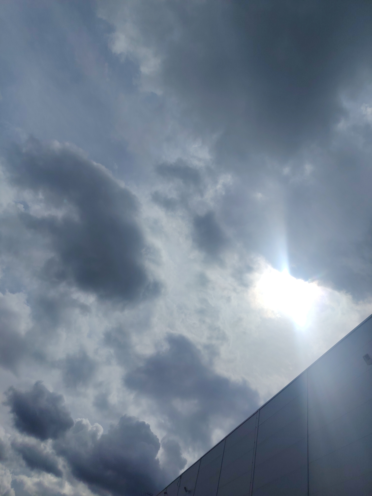 The image depicts a segment of a modern, large-scale building with a light-colored, panelized facade, angled diagonally from the bottom right corner towards the top left. Above the building, a dramatic sky is visible, dominated by dark, heavy storm clouds. Sunlight breaks through a gap in the clouds on the right side of the frame, casting a bright glare and visible rays. The overall atmosphere is brooding and overcast, with a hint of sunlight piercing the gloom. The location is identified as Хоругвино, Russia. There are no people or specific actions discernible in the image.
