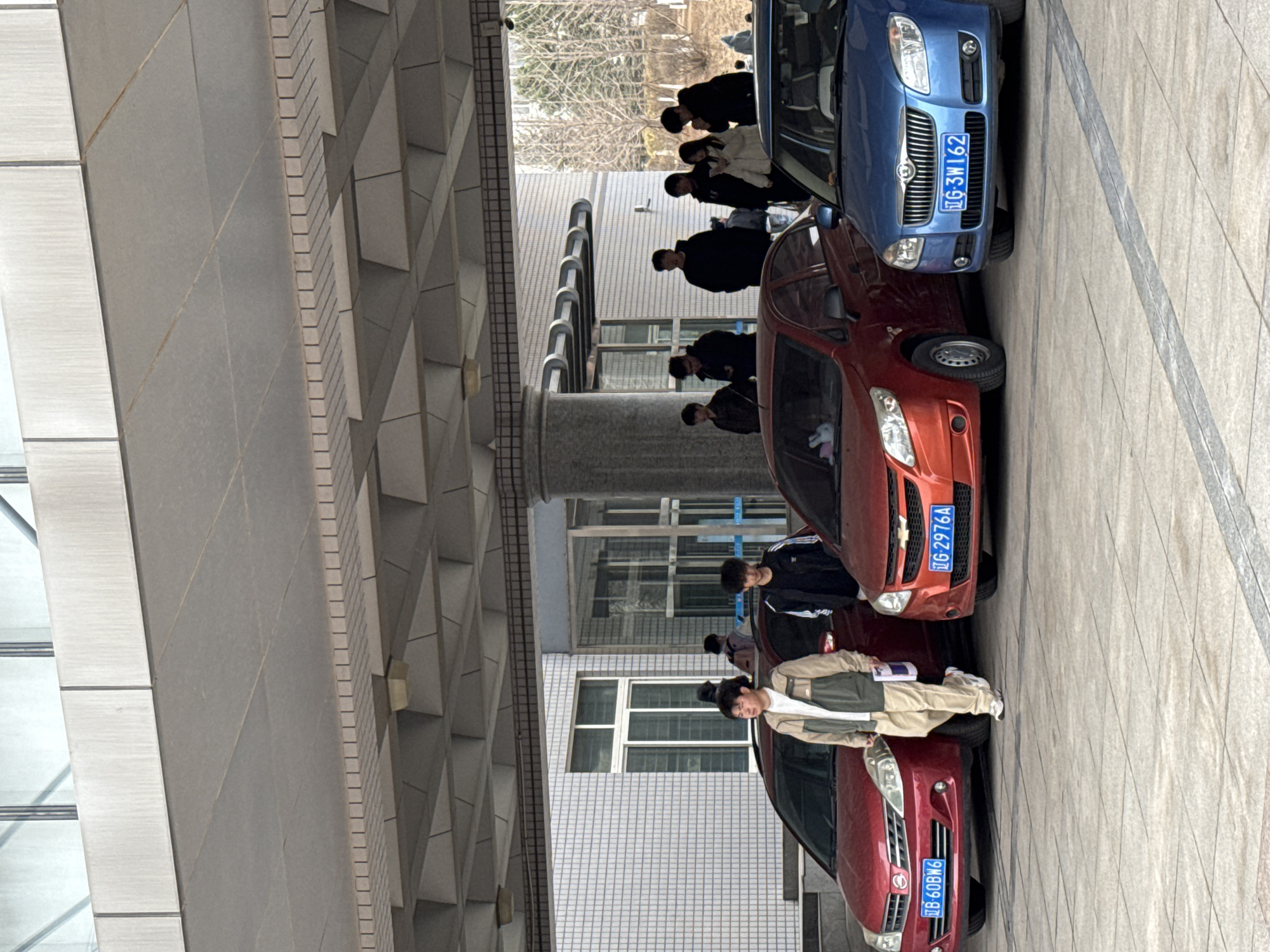 The image depicts a street scene in Zhongtun, China, featuring pedestrians and several parked cars. People are walking along the sidewalk. A blue car with license plate "TG-3W162", a red car with license plate "TG-2976A", and another red car with license plate "B 60BN6" are visible. The environment includes a building with a distinctive geometric overhang. The street is paved with patterned tiles. The overall scene appears to be an ordinary street with typical urban activity.