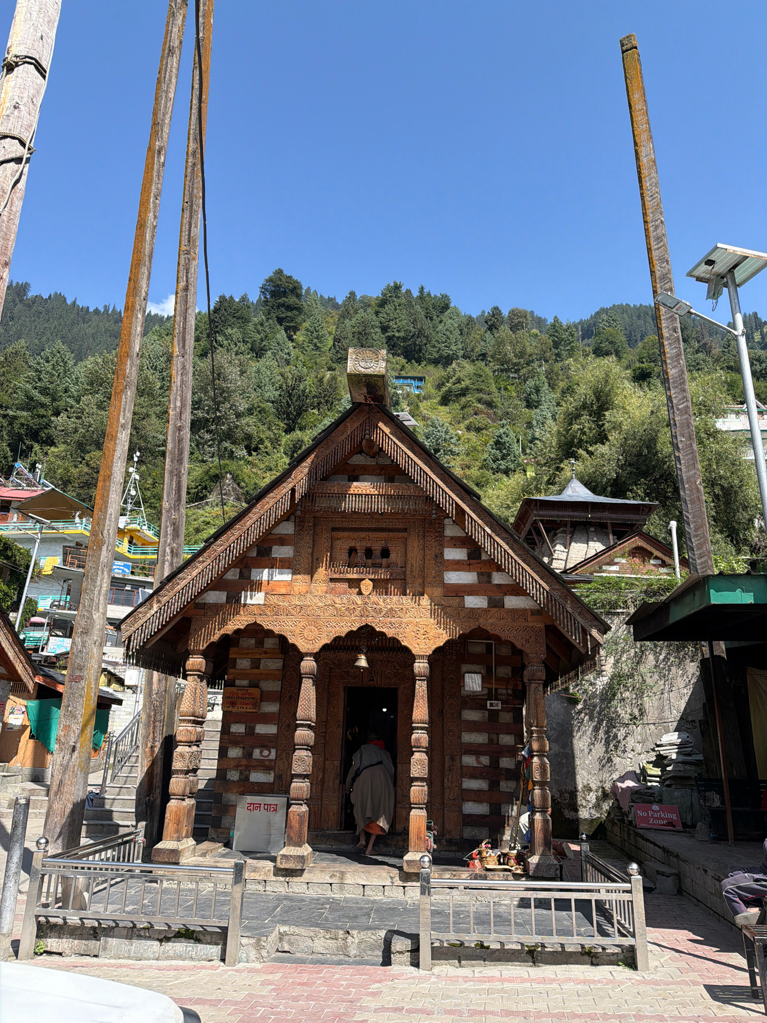 The image captures the entrance of a wooden temple in Vashisht, India, likely a Hindu temple given the architectural style and the "दान पात्र" (donation box) sign. The temple features intricate wood carvings on its facade, a steeply pitched roof, and carved wooden pillars supporting the entrance. A person in a long, light-colored robe is entering the temple from the right. The temple is situated on a hillside covered with dense green trees. In the foreground, there are two tall wooden poles, one on the left and another on the right, which appear to be part of the temple grounds or a local tradition. A solar-powered street lamp is visible on the right pole. A "No Parking Zone" sign is posted on a wall to the right of the temple entrance. The ground in front of the temple is paved with red and grey bricks, and a metal railing encloses the entrance area. The sky is clear and blue. The scene suggests a sunny day in a mountainous region of India, likely during daytime.