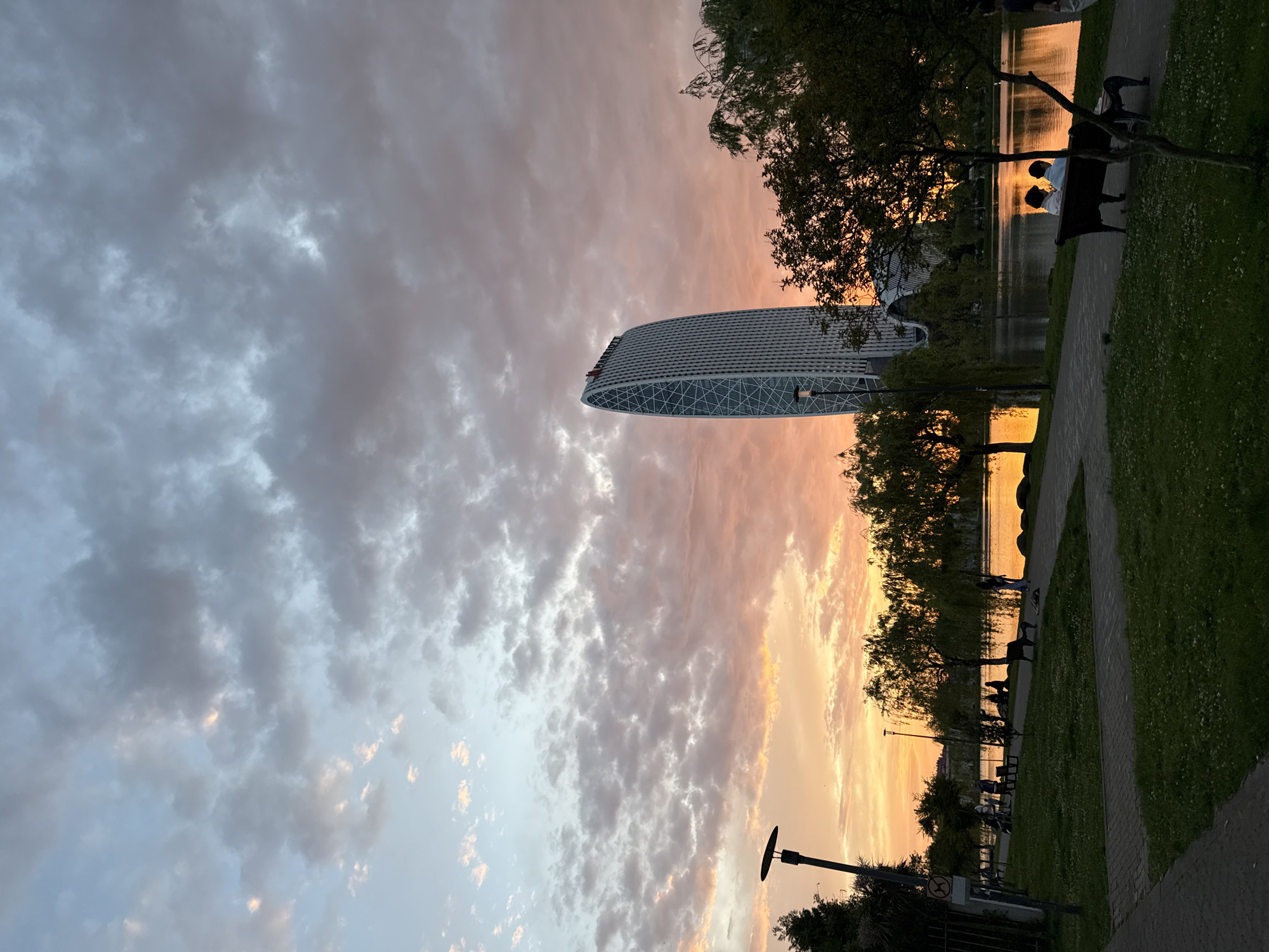 The image depicts a sunset scene in Batumi, Georgia. A modern, elliptical skyscraper stands tall amidst trees and a park area. A body of water, likely a lake or the sea, reflects the orange and yellow hues of the setting sun. The sky is filled with clouds that are tinted with the colors of the sunset. Along the park's edge, there are benches and people enjoying the view. A paved pathway runs alongside the water. A lamppost is visible near the bottom edge of the image.