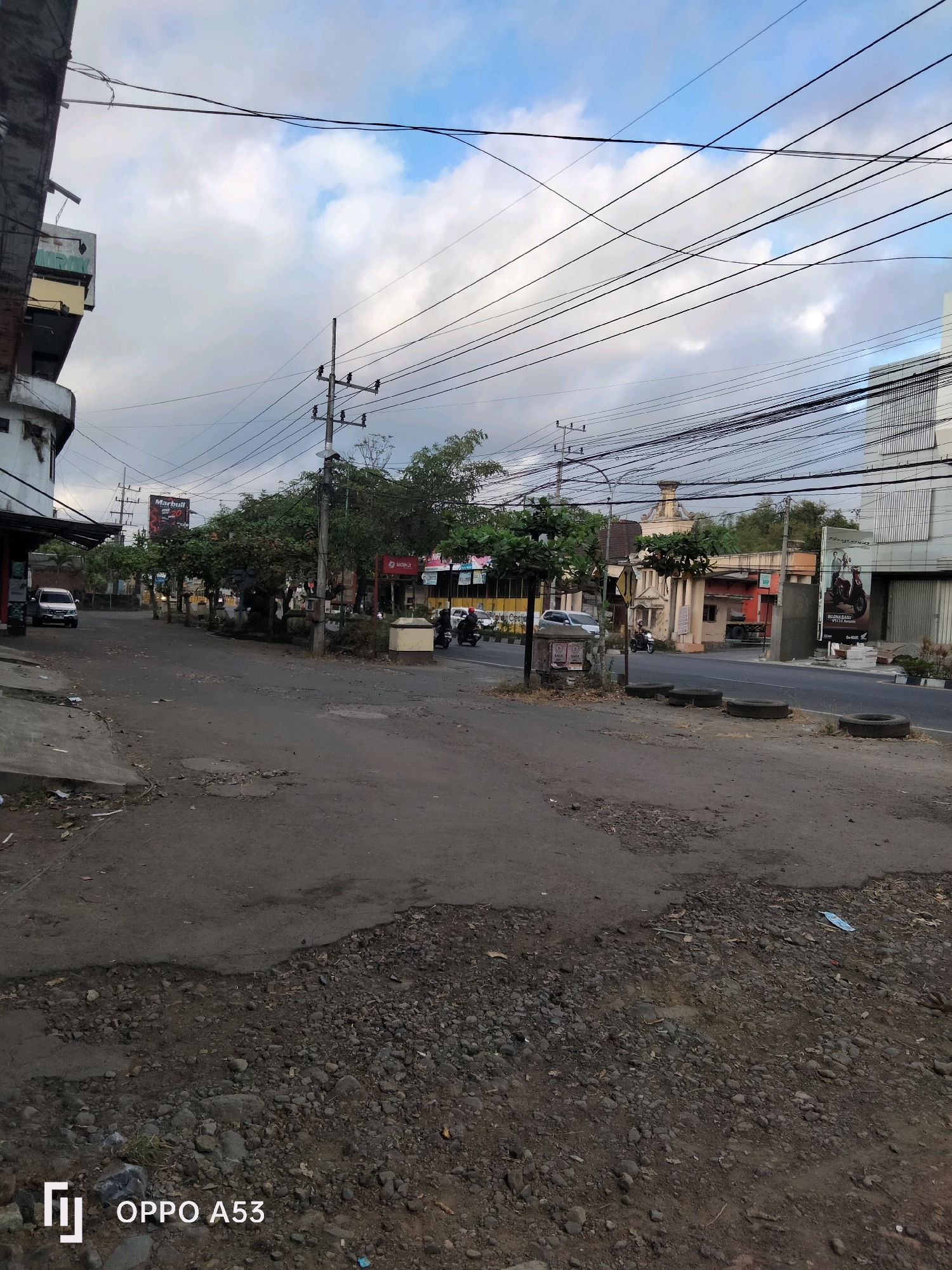 The image depicts a street scene in Trenggalek, Indonesia, characterized by a cloudy sky and a somewhat unkempt urban environment. The foreground shows a rough, unpaved surface covered with gravel and debris, contrasting with the asphalt road in the midground. Several vehicles are visible on the road, including cars and motorcycles.
On the left side of the street, a building with a partially obscured facade stands, appearing somewhat weathered. Further down the street, utility poles with a dense network of electrical wires crisscross overhead, a common sight in many Indonesian cities. Trees line the sides of the road, interspersed with buildings and commercial establishments.
In the background, a large billboard advertising "Marbl" is prominently displayed, suggesting commercial activity in the area. Other buildings, including a structure with a distinctive arched entrance, are also visible. The overall impression is a busy, functional street in a developing urban setting, with signs of daily life and commerce. The presence of the "OPPO A53" watermark in the lower left corner indicates the image was captured with an OPPO A53 smartphone.