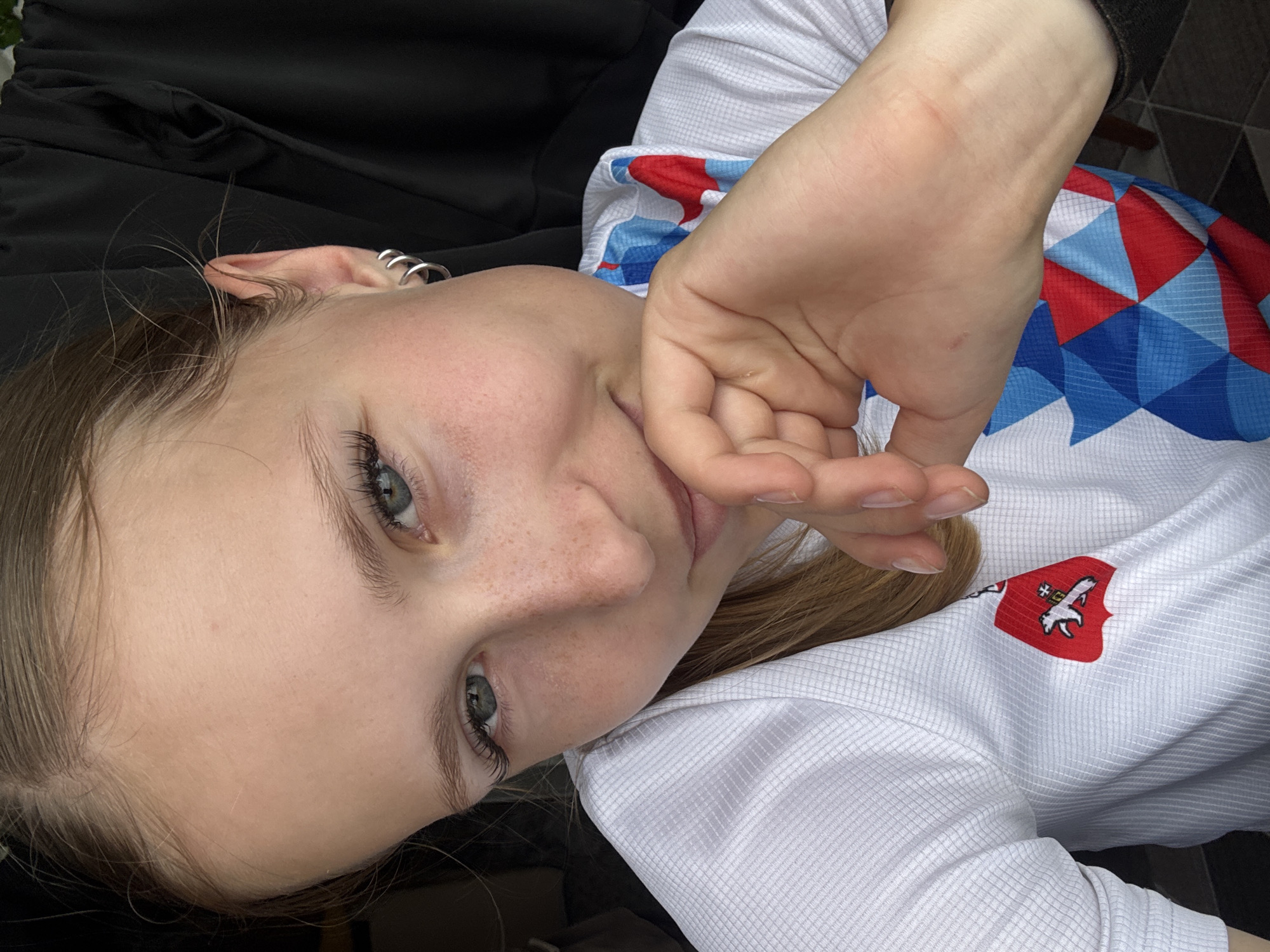 The media file is a close-up, rotated image of a young person's face and upper torso. The subject has light skin with freckles, blue-green eyes, and light brown hair. They are wearing a white t-shirt with a red, blue, and white geometric pattern on the collar and a crest with a white eagle-like figure on a red background on the left side of the chest. The person is looking towards the camera and has their right hand touching their mouth. The background is blurred and appears to be dark fabric, possibly clothing, and a tiled surface. The lighting suggests it could be daytime. The image is oriented sideways. No visible text is present. The context provided indicates the location is Chaikovsky, Russia, but this information cannot be deduced from the image itself.