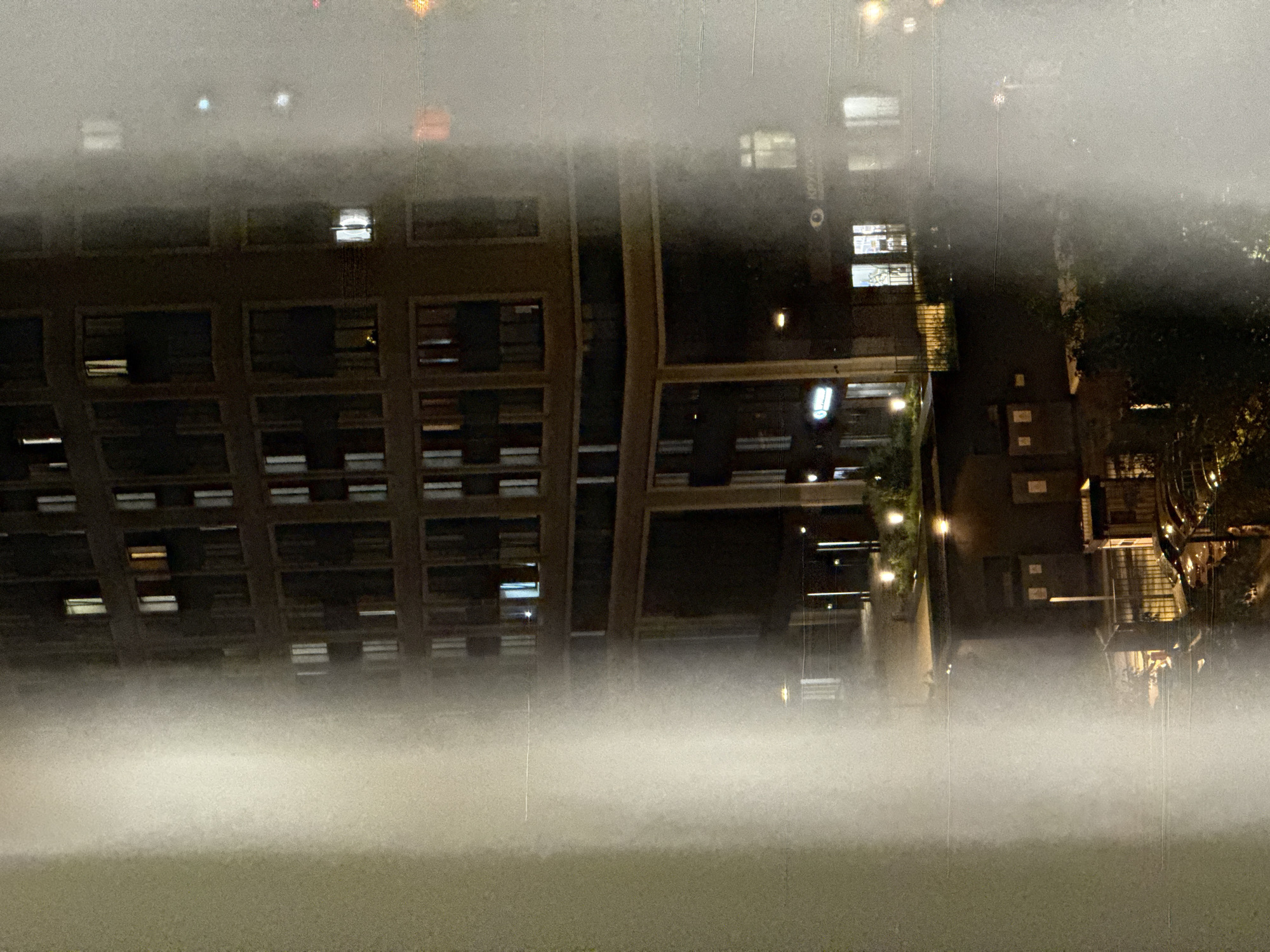 An outdoor photograph, taken at night with low visibility, shows what appear to be buildings and foliage. The perspective is unusual; it's unclear if the image is a reflection, a distorted view, or if the camera was oriented atypically. A heavy fog or mist obscures the lower portion of the view. Several windows are illuminated in the dark buildings, indicating occupancy. Vegetation is evident in the vicinity of the buildings, although the poor lighting limits clarity. The photograph's details are insufficient to confirm the location as Khimki, Russia, or to provide more forensic or specific contextual information. The lack of details also doesn't allow to confirm people, subjects, objects, or actions or interactions.