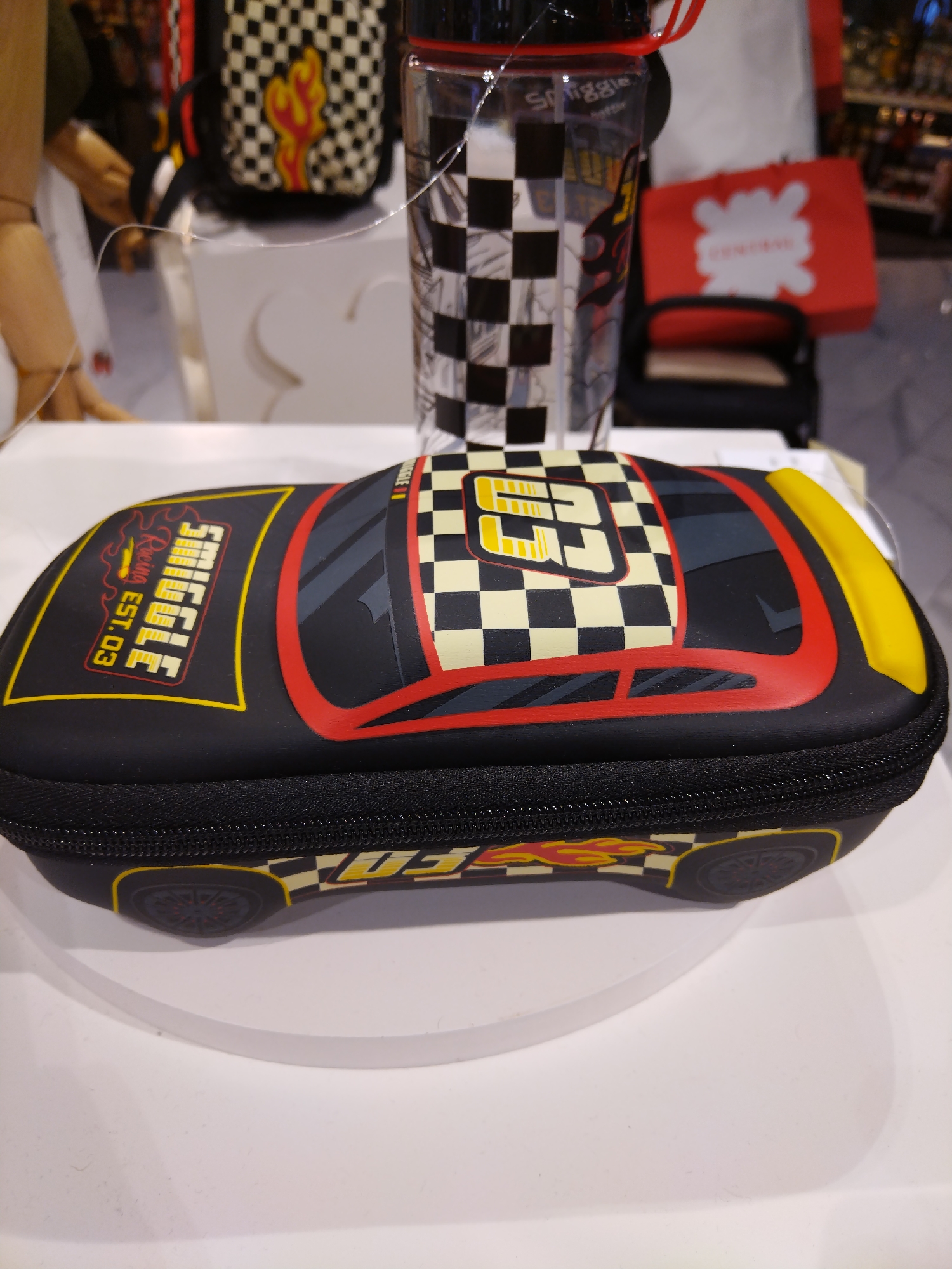 A black zippered pencil case, shaped like a race car with checkered flag patterns, flames, and the word "Smiggle" in red and yellow, sits on a white circular display stand. The pencil case has red trim outlining the car's shape and a yellow spoiler on the back. The background is out of focus, showing a retail environment with shelves and other merchandise, including a clear plastic bottle with a checkered pattern and a red shopping bag. The scene appears to be in a store in Patong, Thailand, given the context.