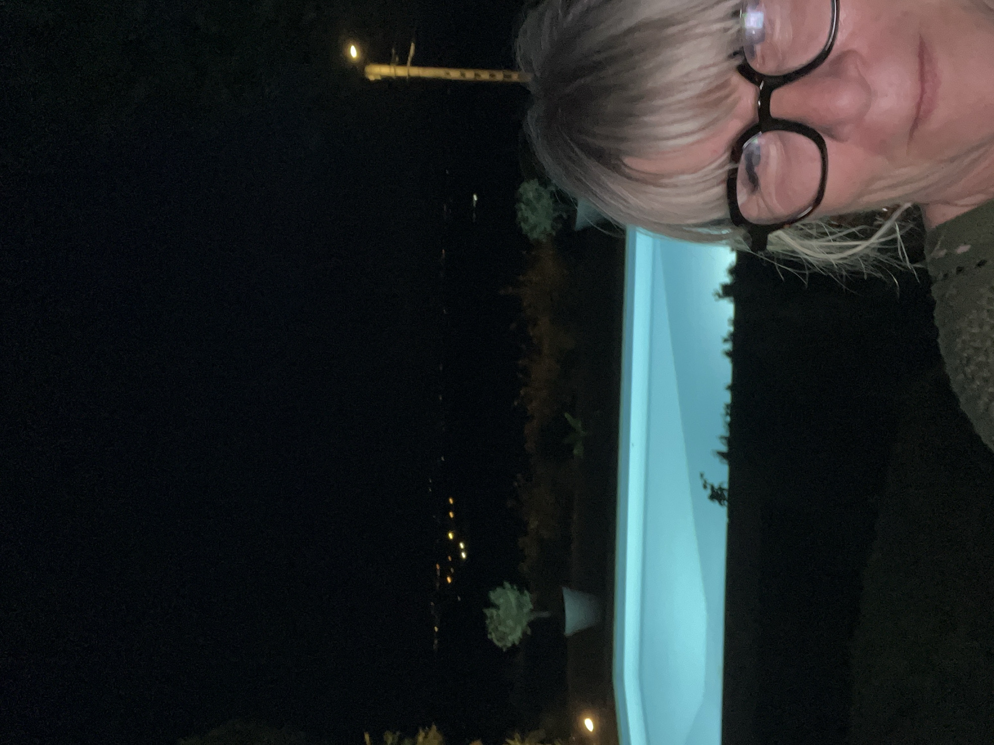 The image shows a nighttime scene with a woman in the foreground wearing glasses. Behind her is a bright blue swimming pool, and beyond that, a dark landscape with distant lights that suggest a town or city. There are also trees and a lamppost visible. The overall impression is that of a residential area in or near Saint-Galmier, France, at night, with the swimming pool illuminated.