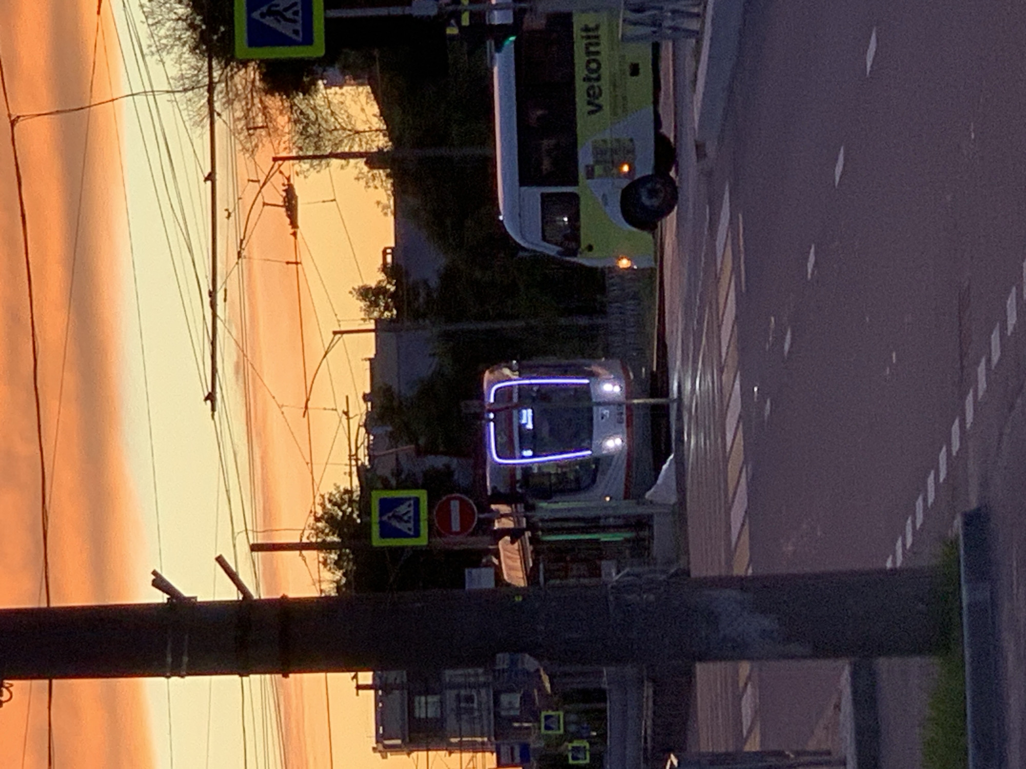 The image captures an urban street scene during either sunrise or sunset, indicated by the prominent orange and yellow hues dominating the sky on the left. The lighting is low, giving the scene a twilight or dawn atmosphere.
In the foreground, a dark utility pole stands vertically on the left. Several overhead power lines, likely for the tram system, crisscross the frame against the bright sky.
The main subjects are two vehicles on a multi-lane road. A modern tram, white with prominent blue and white illuminated lights on its front, is positioned centrally, facing forward. To the right of the tram, a light yellow-green and white bus or van is visible, also facing forward. The word "vetonit" is clearly visible in dark lettering on the side of this vehicle.
Road markings include a visible pedestrian crossing (zebra crossing) and dashed lane lines on the dark asphalt surface. Several traffic signs are present: a blue square sign with a white triangle and a walking person symbol (indicating a pedestrian crossing) is seen above and behind the tram. Another red circular sign with a white horizontal bar (no entry) is located closer to the tram. Buildings and trees are visible in the darker midground and background, silhouetted against the sky or dimly lit by artificial light. The overall impression is an active streetscape transitioning between day and night.