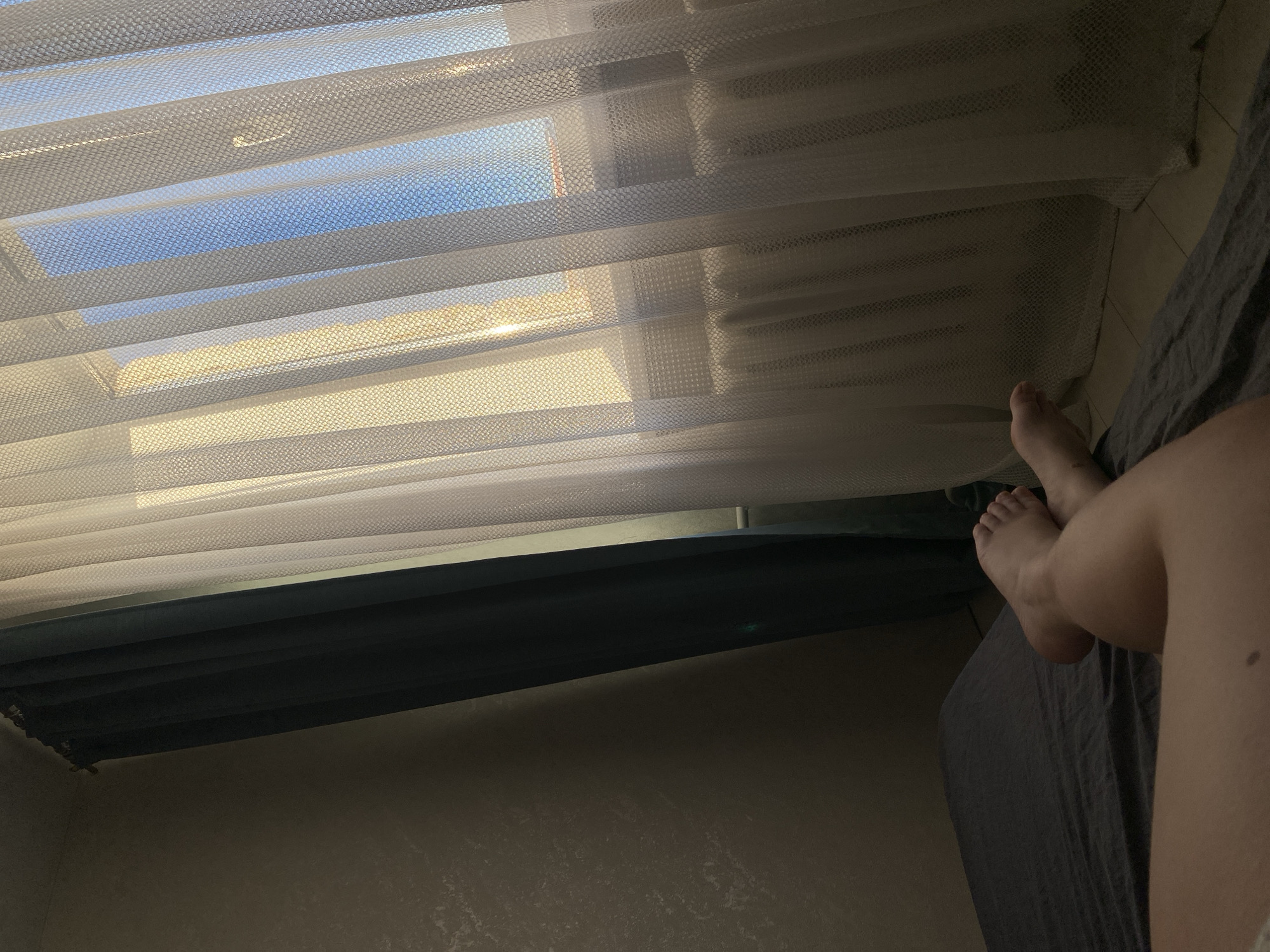 The scene is indoors, viewed from a low angle, possibly from a bed. Light filters through a white, semi-transparent window covering, revealing glimpses of a blue sky and exterior structures. A dark-colored window frame is visible beneath the covering. To the right, a person's legs and feet are partially visible resting on a dark-colored bed covering. The background is a wall. The location is unknown.