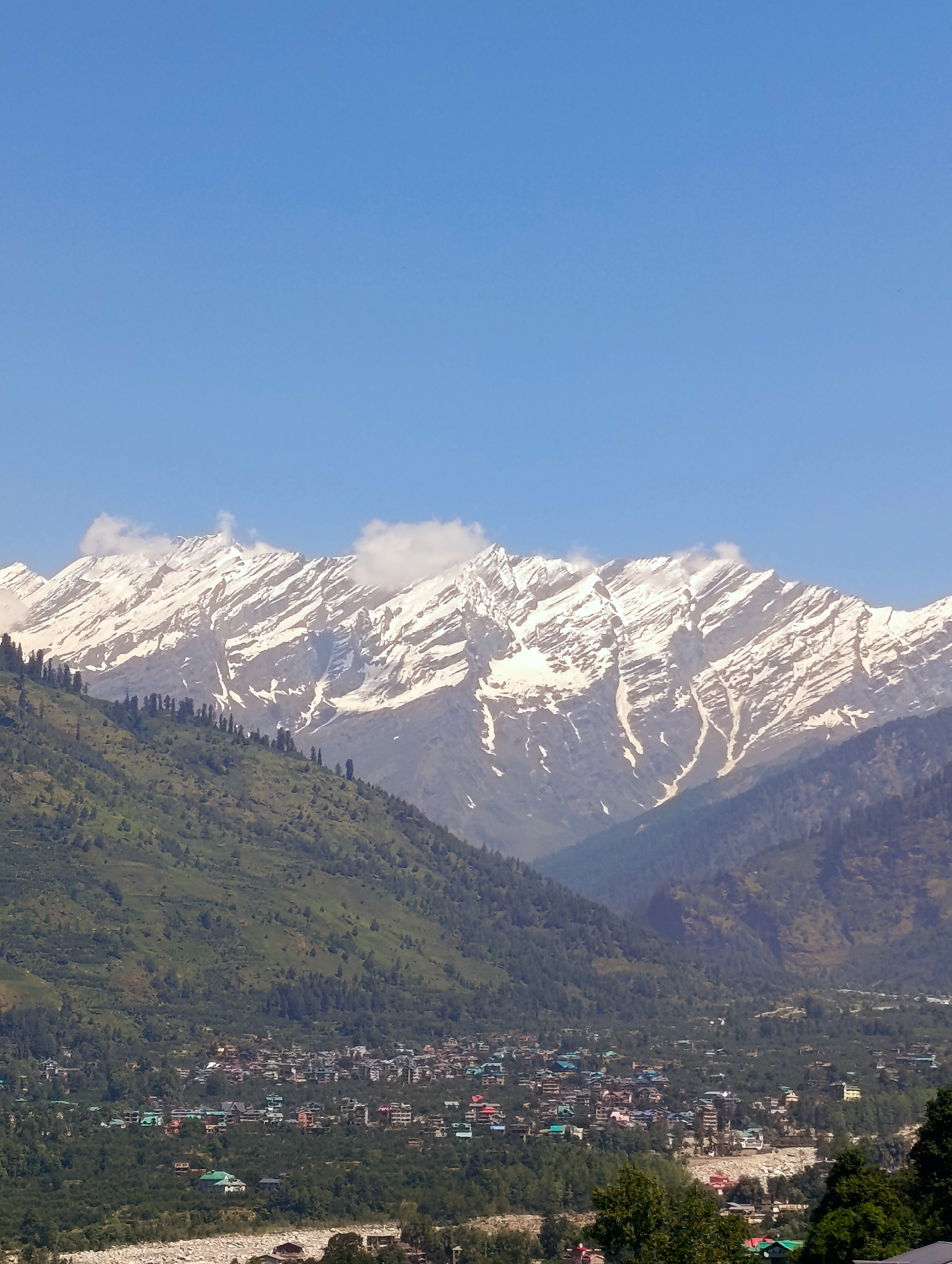 The image depicts a wide panoramic view of a mountain valley with a village nestled within it. In the background, a majestic mountain range is capped with snow, its slopes striated with shadows and lighter patches indicating the snow cover. The sky above is a clear, vibrant blue. In the mid-ground, a lush green mountainside slopes down towards the valley, dotted with scattered trees. The foreground reveals a densely populated village, characterized by numerous buildings with colorful roofs, clustered together amongst more trees. A dry riverbed, filled with white stones, is visible at the very bottom of the frame, with a few buildings situated along its edge. The overall scene suggests a tranquil, scenic location, likely in a mountainous region. The description of Vashisht, India, provides geographical context, placing this scene within a specific location known for its natural beauty and proximity to the Himalayas.
