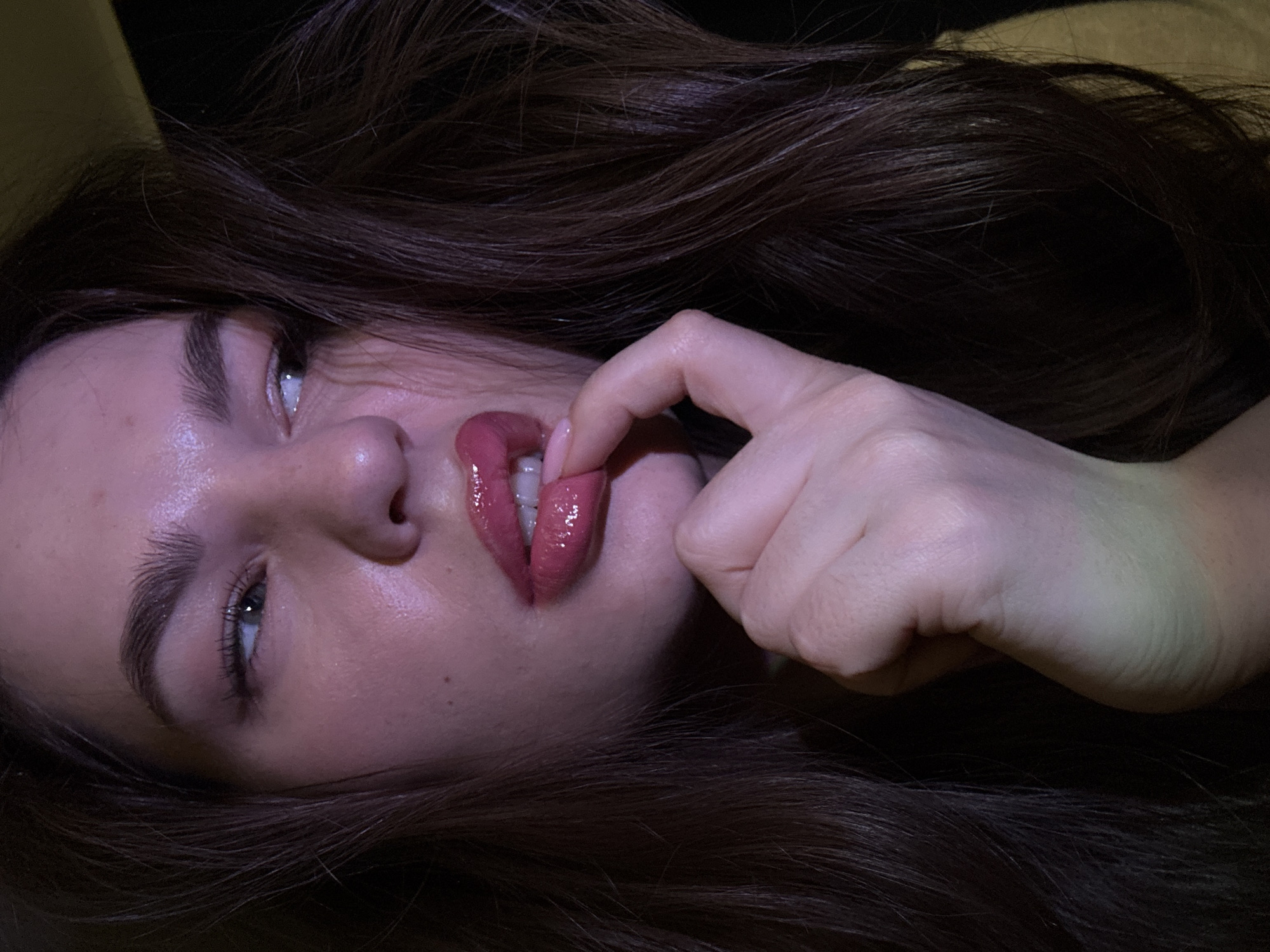 A close-up, angled portrait depicts a woman with long, dark brown hair. She is looking upwards with her left eye visible, which is light blue. Her skin is pale and appears glossy. She has prominent eyebrows and eyelashes. Her lips are full, covered in a glossy pink lipstick, and slightly parted. Her right index finger is touching her lower lip. The background is dark and indistinct. There is no information to indicate that the location is Chaikovsky, Russia.