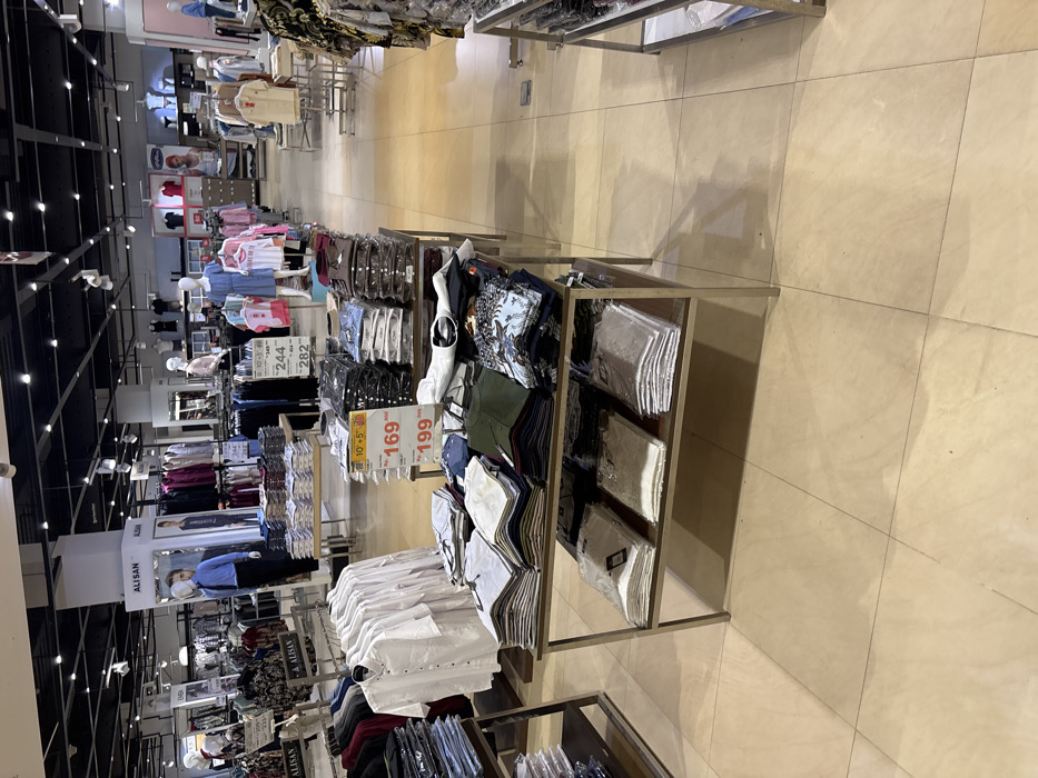 The image shows the interior of a clothing store in a shopping mall located at the coordinates -7.44 latitude and 112.715 longitude in the city of Kecamatan Sidoarjo, Indonesia. The store is well-lit with a tiled floor. The image shows the clothes displayed on two metal shelves with wooden tops. The shelves are positioned near the center of the image. The left shelf is full of neatly folded shirts and the right shelf is full of folded t-shirts. A yellow price tag for 169,000 and 199,000 rupiah is hanging on the left shelf. The right shelf is empty except for stacked t-shirts. In the background, there are shelves full of clothing, and mannequins displaying various items. The store's lighting is overhead, the ceiling is black with a grid pattern and the floor is a light tan.