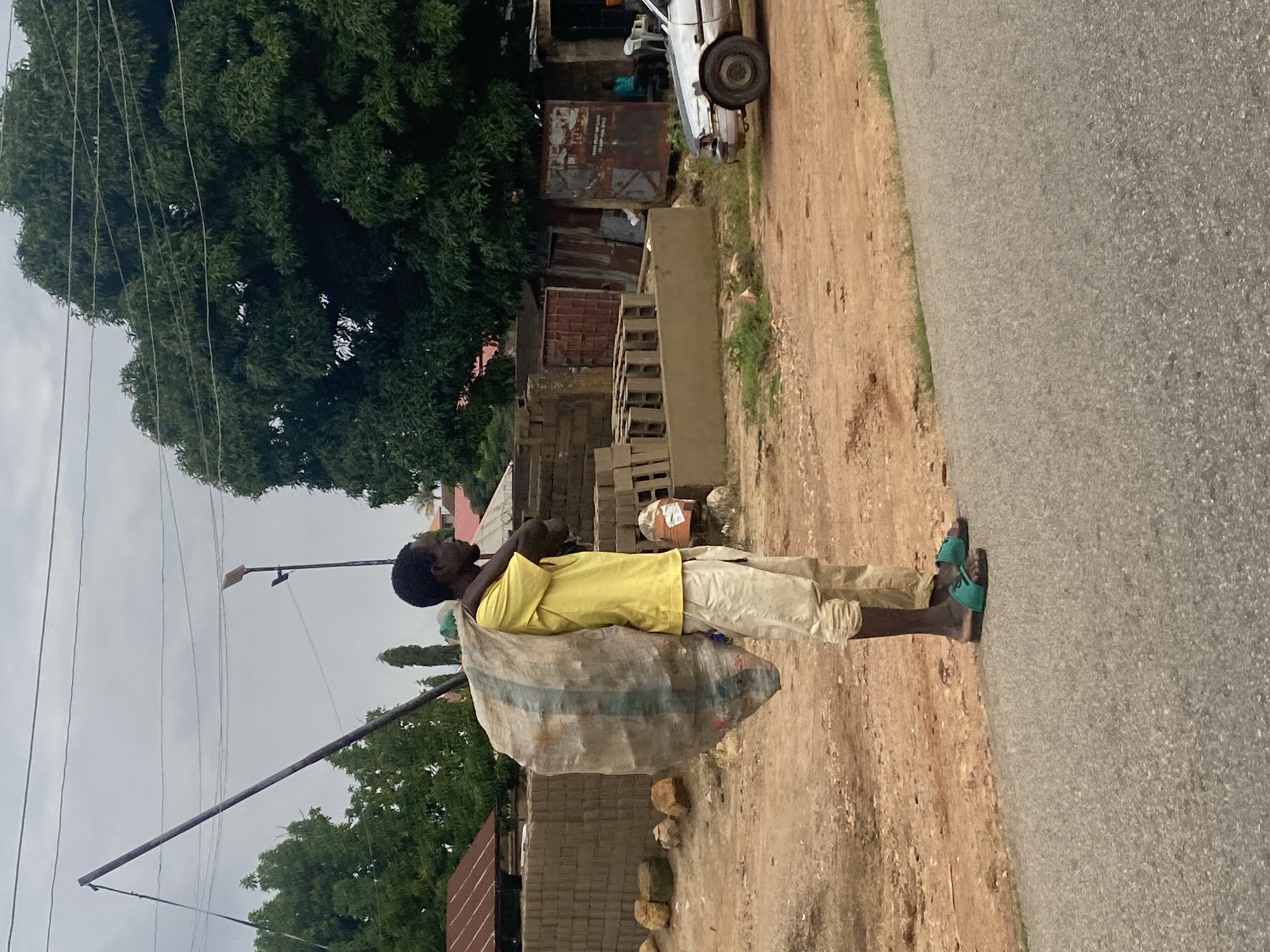A man, likely of West African descent, stands on a dirt road in Jos, Nigeria, carrying a large, transparent plastic bag filled with materials slung over his left shoulder. He is wearing a bright yellow t-shirt and light-colored, loose-fitting trousers that are rolled up at the bottom. He also wears green flip-flops.
The environment suggests an urban or semi-urban setting. Behind him, stacks of what appear to be bricks or building materials are visible, along with some rudimentary structures made of corrugated metal and wood. A large, leafy tree dominates the background, and power lines crisscross the sky, common in urban areas. A silver sedan is parked on the right side of the road, and a few individuals are seated near one of the structures. The ground is dusty, with patches of sparse vegetation. The overall impression is one of a working-class area on an overcast day.