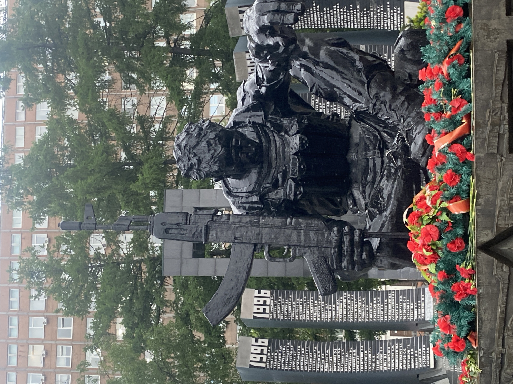 The image captures a somber war memorial, likely located in Yekaterinburg, Russia. The central focus is a dark, possibly bronze or black stone, sculpture of two soldiers, one appearing to support the other. The soldier in the foreground carries an AK-pattern assault rifle. To the right of the sculpture is a memorial wall, decorated with a wreath of red carnations and possibly other greenery, adorned with names inscribed on the wall, most likely those of fallen soldiers. Behind the sculpture and memorial walls are several gray blocks, also covered with inscriptions listing names and the year "1996," suggesting a specific conflict or period. The background consists of a building with multiple stories and numerous windows, partially obscured by green trees. The overall tone is solemn and commemorative, honoring those who served and were lost in war.