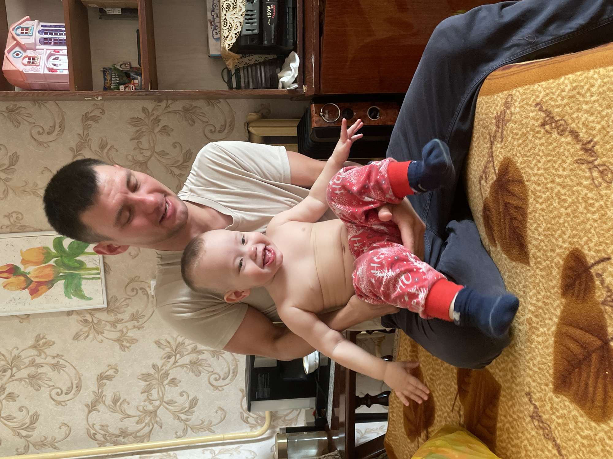 A man is seated, holding a baby in his lap. The baby is wearing red and white pants and dark blue socks. The man is wearing a light-colored t-shirt and dark pants. They are indoors, with patterned wallpaper on the wall. A painting of tulips hangs on the wall behind them. There is a wooden shelf above them with a toy house on it. A brown blanket covers the chair they are seated on. This image was taken in Siva, Russia.