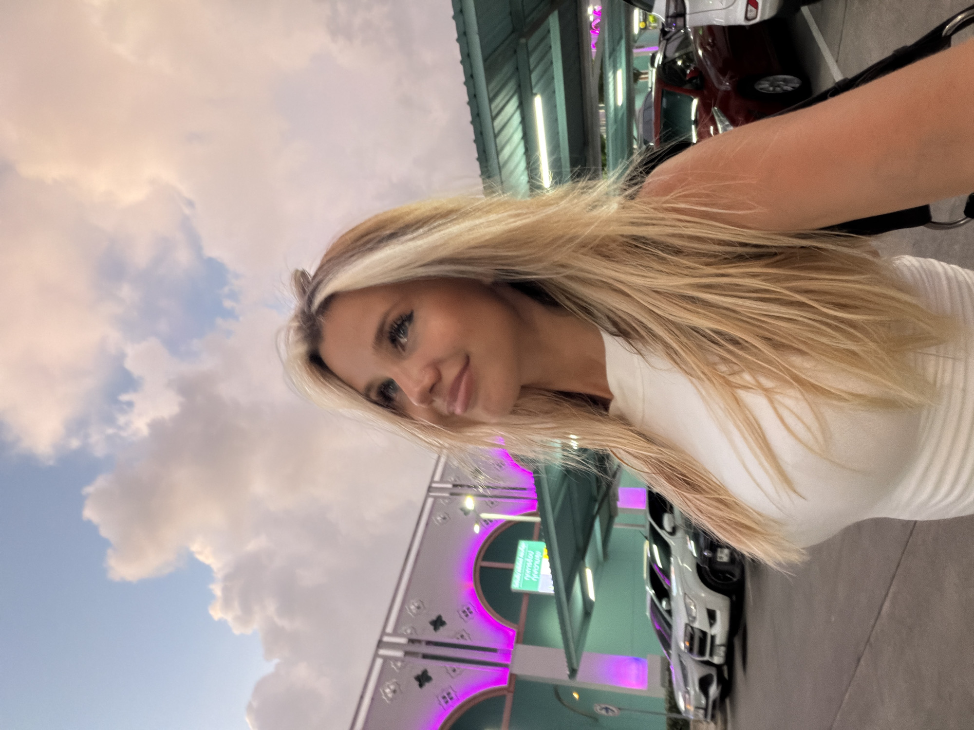 A young Caucasian woman with blonde hair and blue eyes is standing in a parking lot in front of a building with pink lighting. She is wearing a white tank top and looking up and to the right. The sky is cloudy with a mix of blue and pink hues, suggesting it is either sunrise or sunset. Several cars are parked in the lot behind her. The building appears to be a commercial establishment, possibly a shopping mall or entertainment venue, with architectural details that include arched windows and decorative patterns. A sign with Thai text is visible on the building. The overall impression is a casual, outdoor scene in an urban environment in Si Sunthon, Thailand.