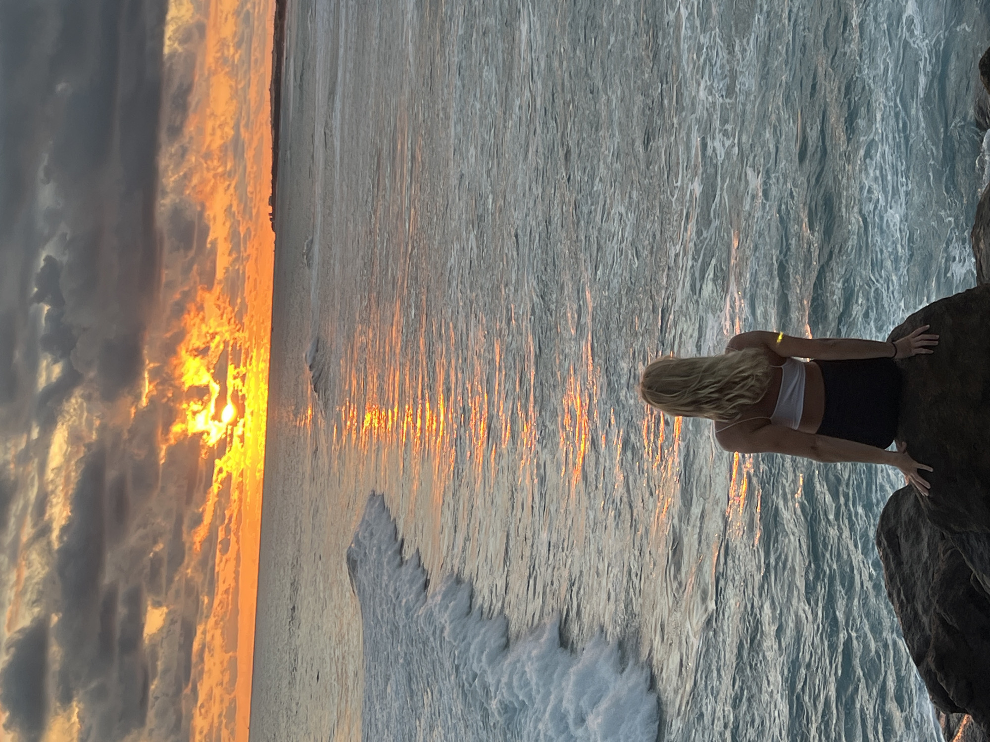 A long shot shows a blonde woman in a white top and dark shorts standing on a rock outcrop on the beach at sunset. The woman's back is to the camera. The sky is a mix of gray clouds and orange sunset. The ocean reflects the orange sunset. The waves are small. The location is likely Goviyapana, Sri Lanka.