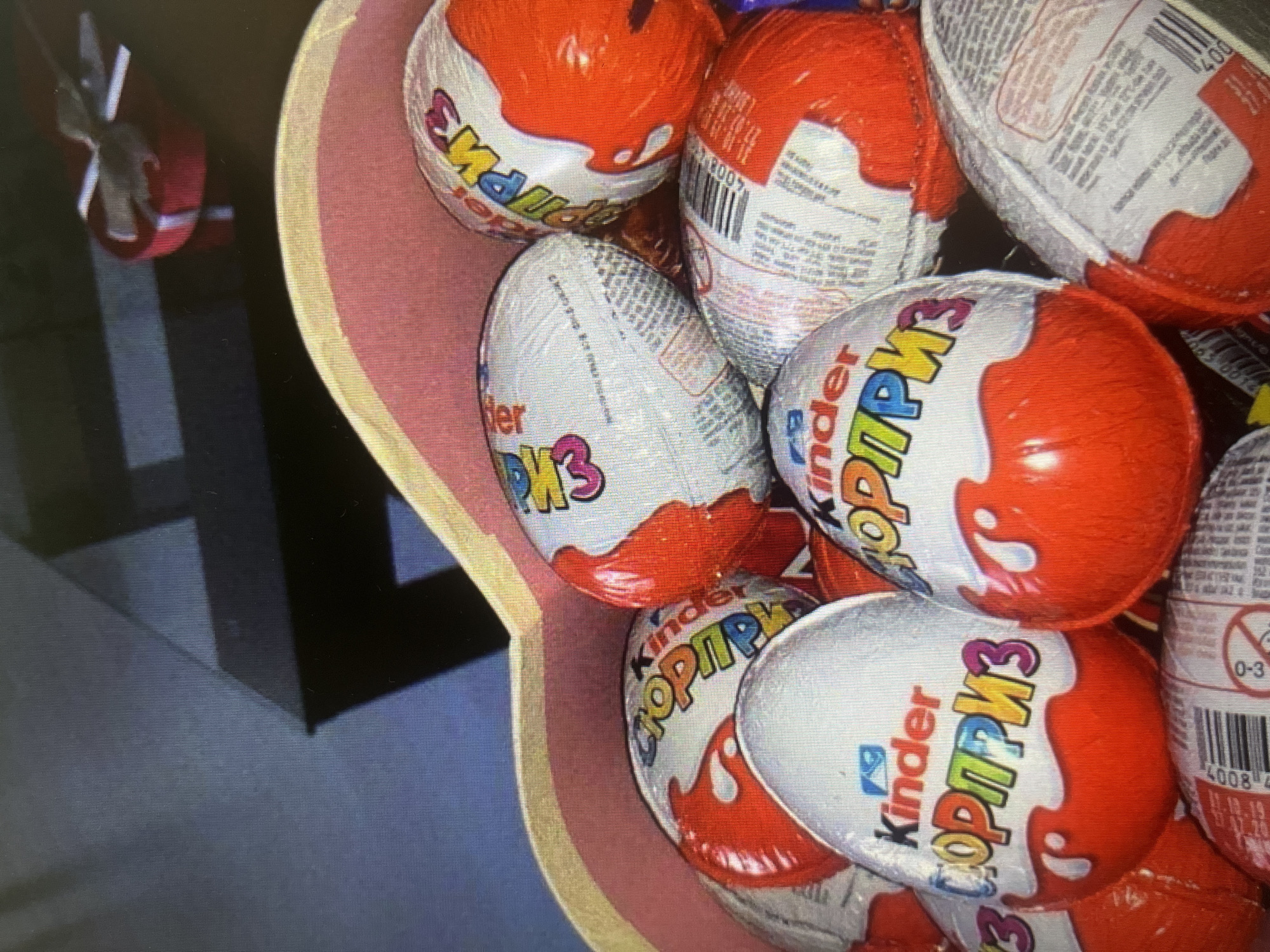 In a close-up, slightly blurry photograph, a collection of Kinder Surprise eggs are densely packed within a pink and yellow heart-shaped container. The eggs are wrapped in their iconic foil, predominantly red and white, with the Kinder logo and Russian text, "СЮРПРИЗ," visible on many of them. Some of the eggs are slightly crushed or partially obscured by others. In the upper right background, a red heart-shaped gift box with a white ribbon is visible, placed on a dark surface. The overall lighting is dim, suggesting an indoor setting, possibly a home or a gift shop. No people or specific locations within Moscow are identifiable in the image.