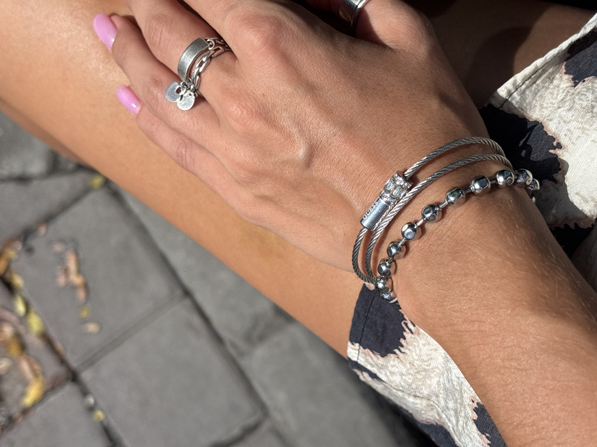 A person's right arm and hand, adorned with silver jewelry, are prominently featured in this medium close-up shot. The jewelry includes two bracelets, one with a series of silver balls and another with a silver cylinder encrusted with small clear stones, and a ring on the index finger. The ring is silver with a chain-like band and a heart-shaped charm. The person's fingernails are painted a bright pink. The background consists of gray paving stones with scattered yellow and brown leaves, suggesting an outdoor urban environment. The lighting is bright, casting shadows and highlighting the textures of the skin and jewelry.
