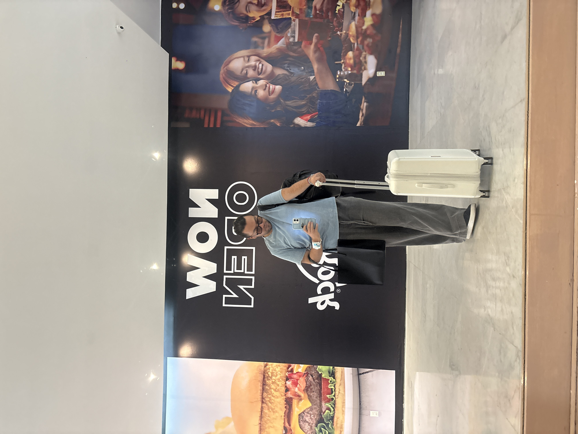 The photograph, taken indoors, depicts a man with a suitcase standing in front of a Hard Rock Cafe advertisement. The man is facing the camera, holding a phone in his hand, and wearing glasses, a light blue shirt, and gray pants. His luggage is a white suitcase with a telescoping handle extended. Beside him is a black shopping bag.
Behind the man is a large advertisement that features a group of people at the top. Beneath them are the reversed words "NOW OPEN," and the "Hard Rock" logo. Below this is a large image of a hamburger.
The walls and floor appear to be constructed of marble-like material. It appears to be taken in Mexico City, Mexico.