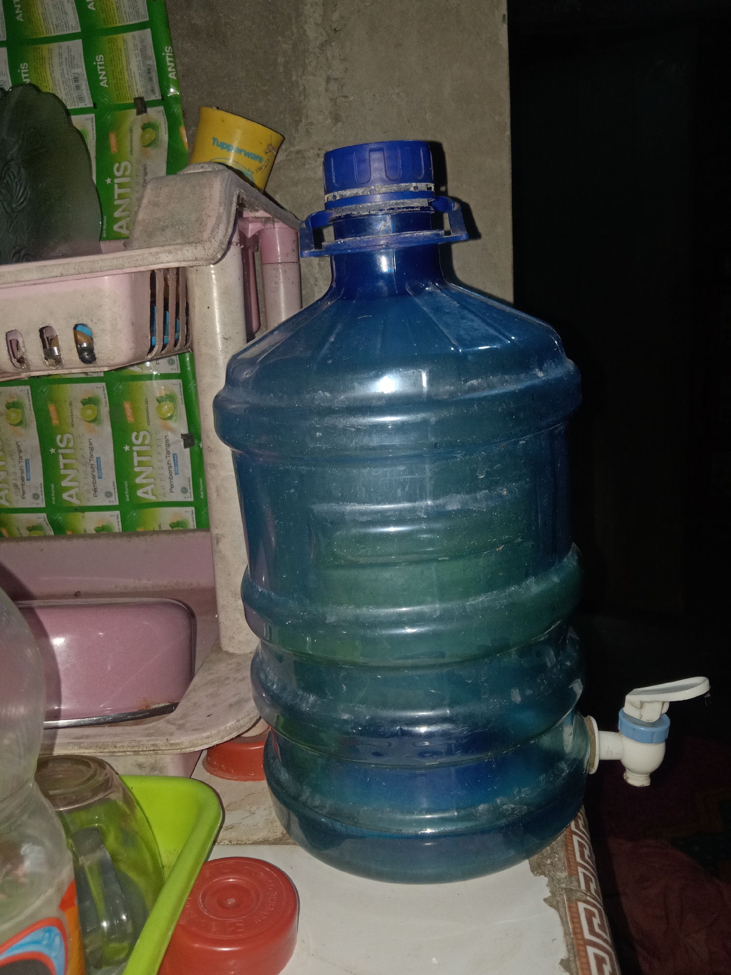 A large, blue, plastic water dispenser with a white and blue tap is centrally located in the foreground. The dispenser is positioned on a tiled surface. To the left of the dispenser, a pink plastic container is visible, and further to the left, a partially visible plastic bottle and a green plastic tray containing a glass are present. Behind the water dispenser, a shelf or tiered structure holds several green and white packaged items. The background is a dark, indistinct wall. The scene appears to be indoors. The image is taken in the city of Jumput, Indonesia.