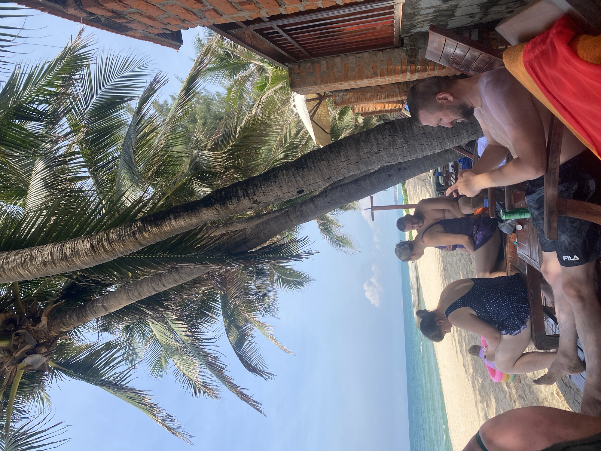 In Phu Thuy Ward, Vietnam, a group of people are relaxing on a beach under the shade of palm trees. The scene is set on a sandy beach next to the ocean, with clear blue water and a bright sky. A brick building with a fence is visible on the right side of the image, suggesting a resort or beachfront property.
Several individuals are seated on lounge chairs. In the foreground, a man with a beard is looking at his phone. To his left, a woman in a polka dot swimsuit is seated, facing away from the camera. Further back, another woman in a purple swimsuit is also seated, with a man behind her. A small inflatable pool is on the sand near the woman in the polka dot swimsuit, and a child can be seen next to it.
Palm trees with their distinctive trunks and fronds dominate the upper portion of the image, providing shade. Beach chairs are arranged on the sand. Various objects are scattered around, including a green can, a bottle, and towels. The overall atmosphere suggests a leisurely day at the beach.