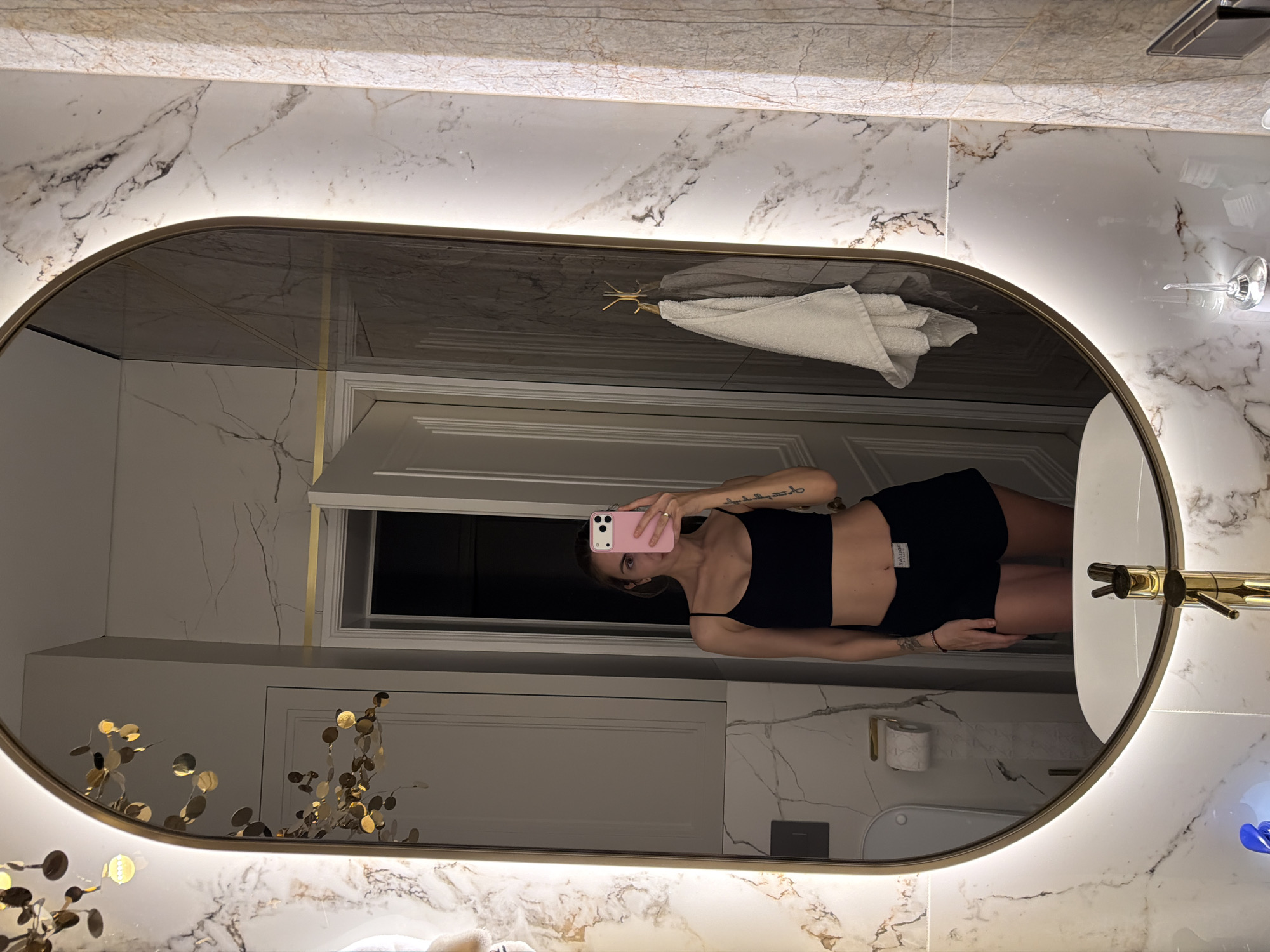 The image displays a mirror selfie taken by a young woman in a modern bathroom, presented rotated 90 degrees clockwise. The woman, who has light skin and a slender physique, is reflected in a prominent oval mirror featuring an illuminated gold frame. She is dressed in a black spaghetti strap crop top and black shorts. Her face is partially obscured by a light pink smartphone that she holds up to take the photograph; her expression appears neutral.
The bathroom is characterized by elegant white marble-patterned walls with striking grey and brown veining. Gold accents are a consistent theme, evident in the mirror's frame, a decorative gold leaf-like element visible in the lower and upper left reflections, and a golden faucet or spout on the right side. Within the mirror's reflection, a white hand towel hangs on a golden hook, and a white paneled door is visible in the background. Outside the mirror on the right, a white light fixture and a glass are seen. Further details in the reflection include a toilet paper holder with a roll and a white toilet. The label on the woman's shorts displays the text "SKORELOVE" (inverted). The overall setting conveys a luxurious and well-lit bathroom environment, possibly located in Krasnoyarsk, Russia.